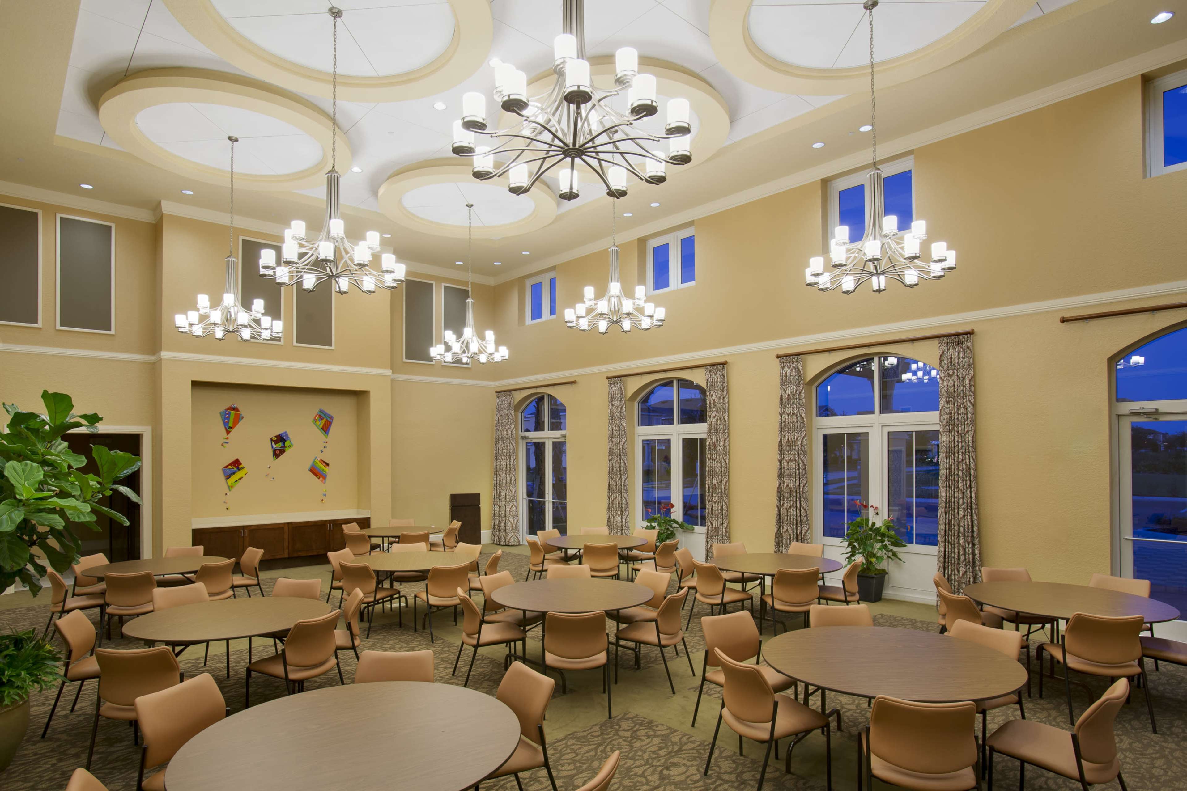 The image shows a spacious indoor dining area with round tables, elegant chandeliers, and large windows allowing ambient light.