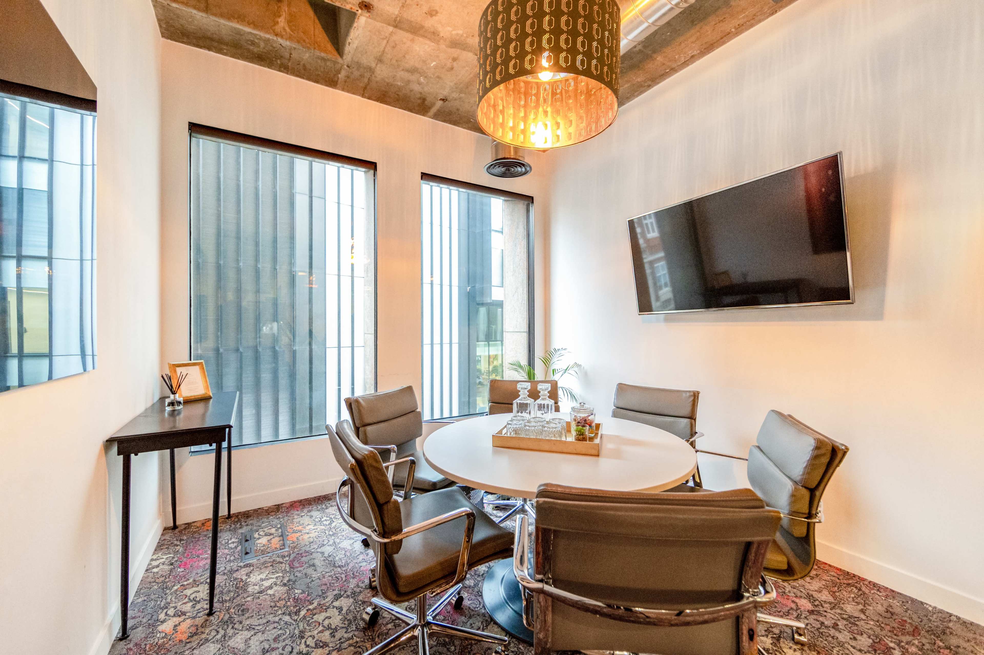 A modern conference room features a round table surrounded by six chairs, a wall-mounted TV, and large windows with a view of the exterior.