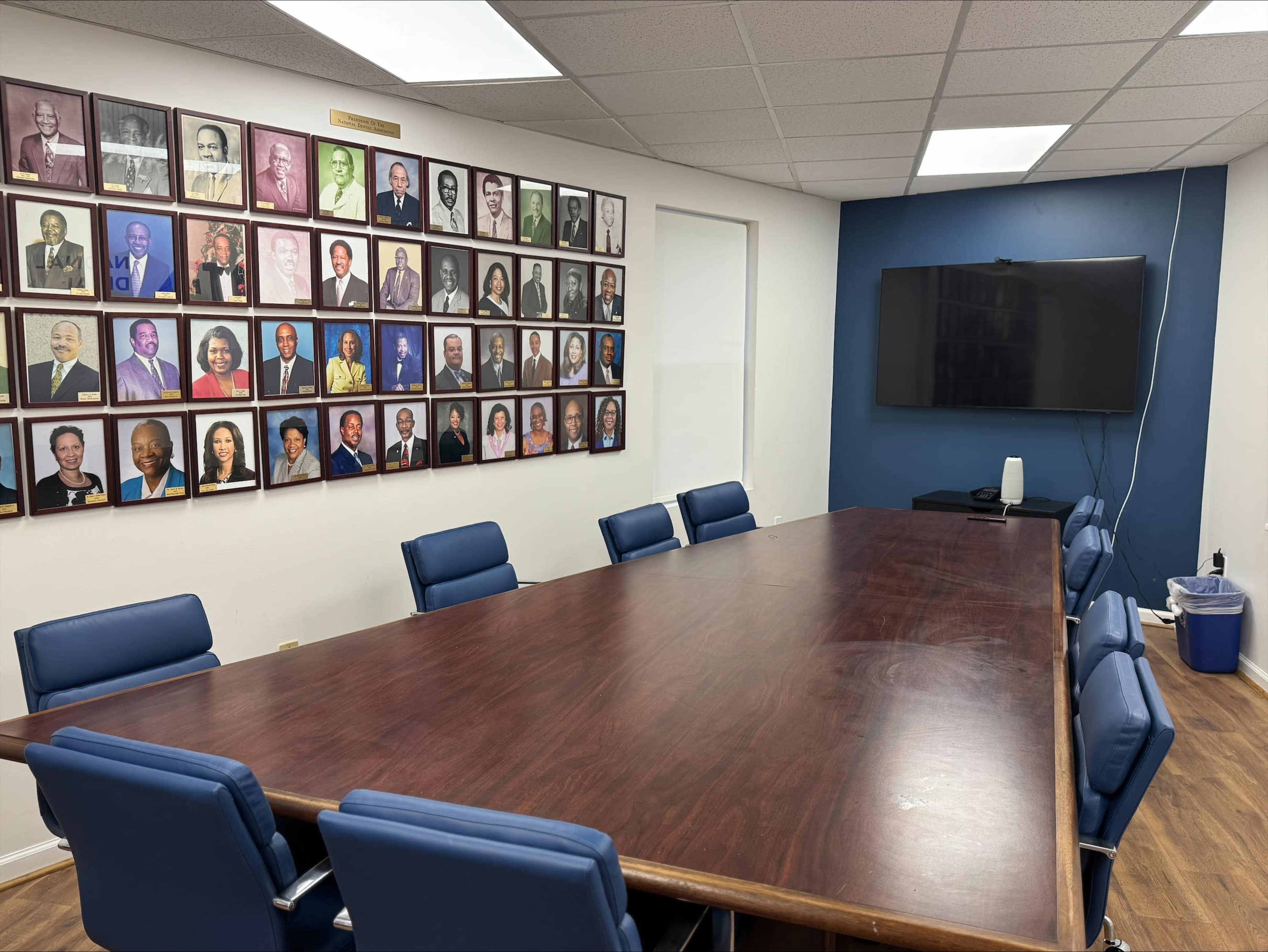 The image shows a conference room with a large wooden table, blue chairs, and a wall featuring framed portraits of various individuals alongside a television mounted on the opposite wall.