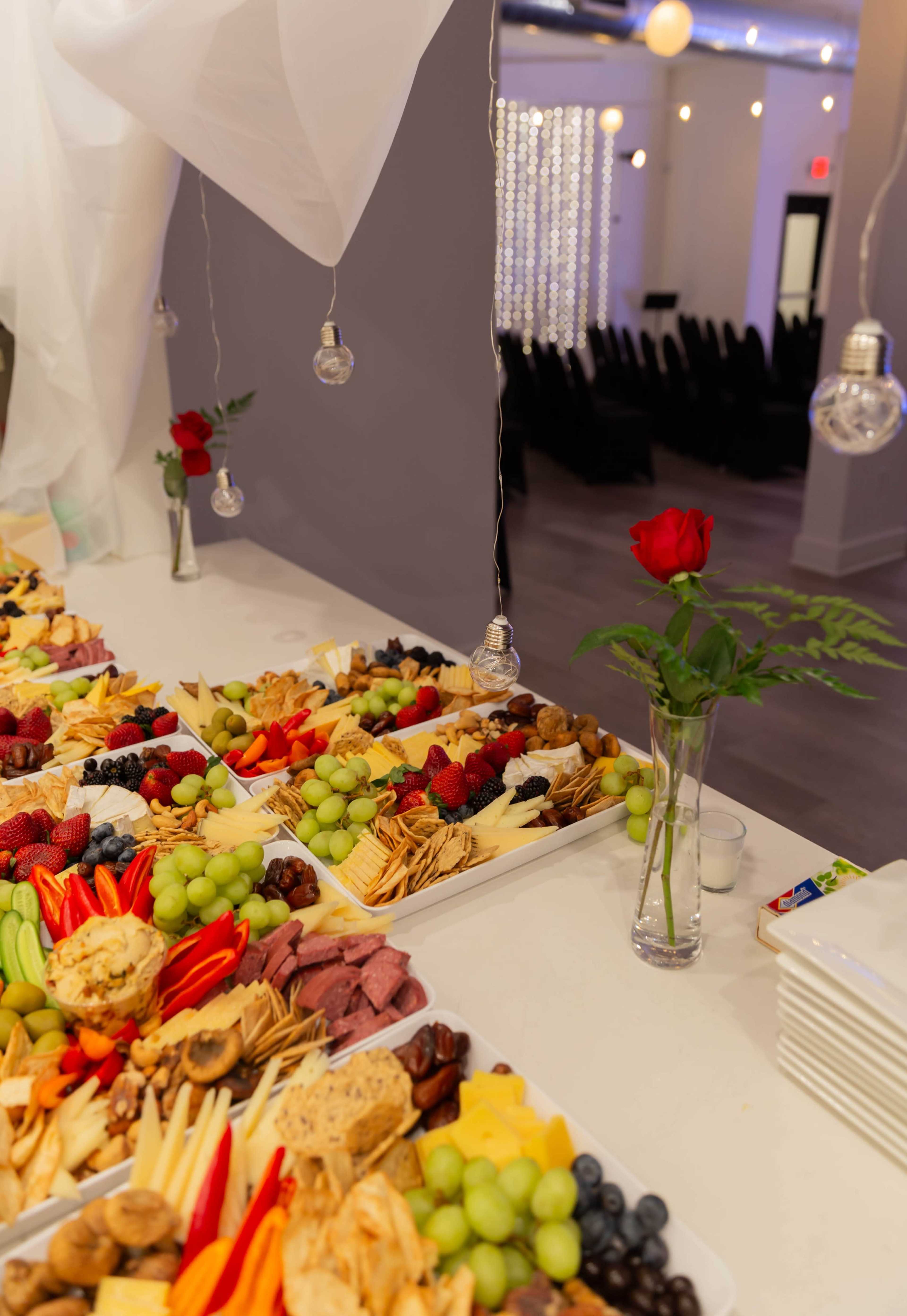 A table features an elaborate display of various cheeses, meats, fruits, and vegetables arranged on platters, with a rose in a vase and decorative lighting in the background.