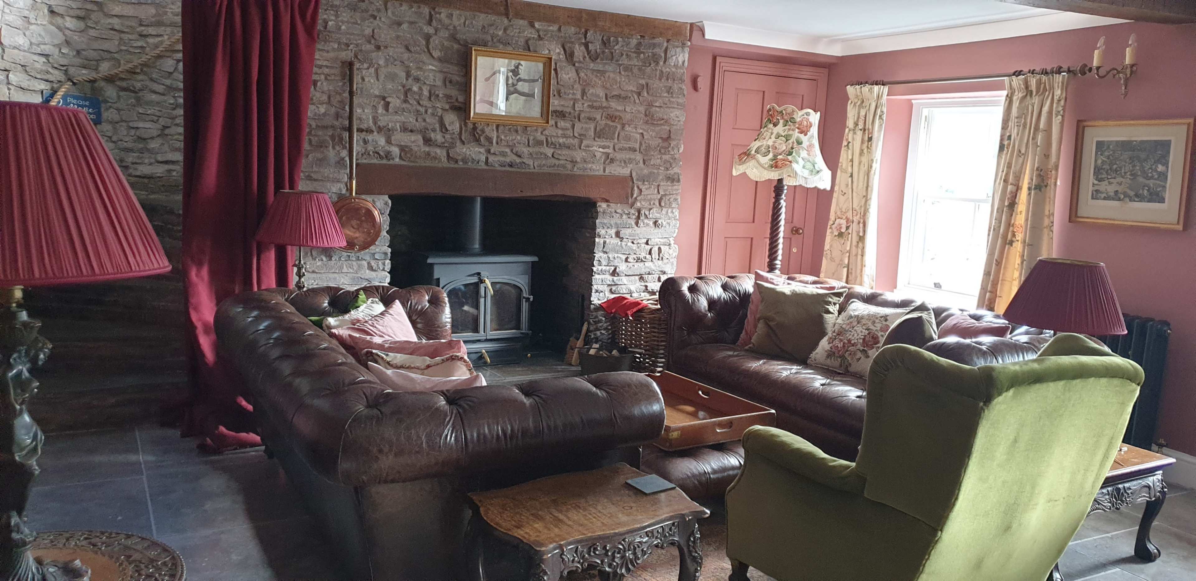 A cozy living room with two brown leather sofas and a green armchair, a stone fireplace, and large windows with curtains.