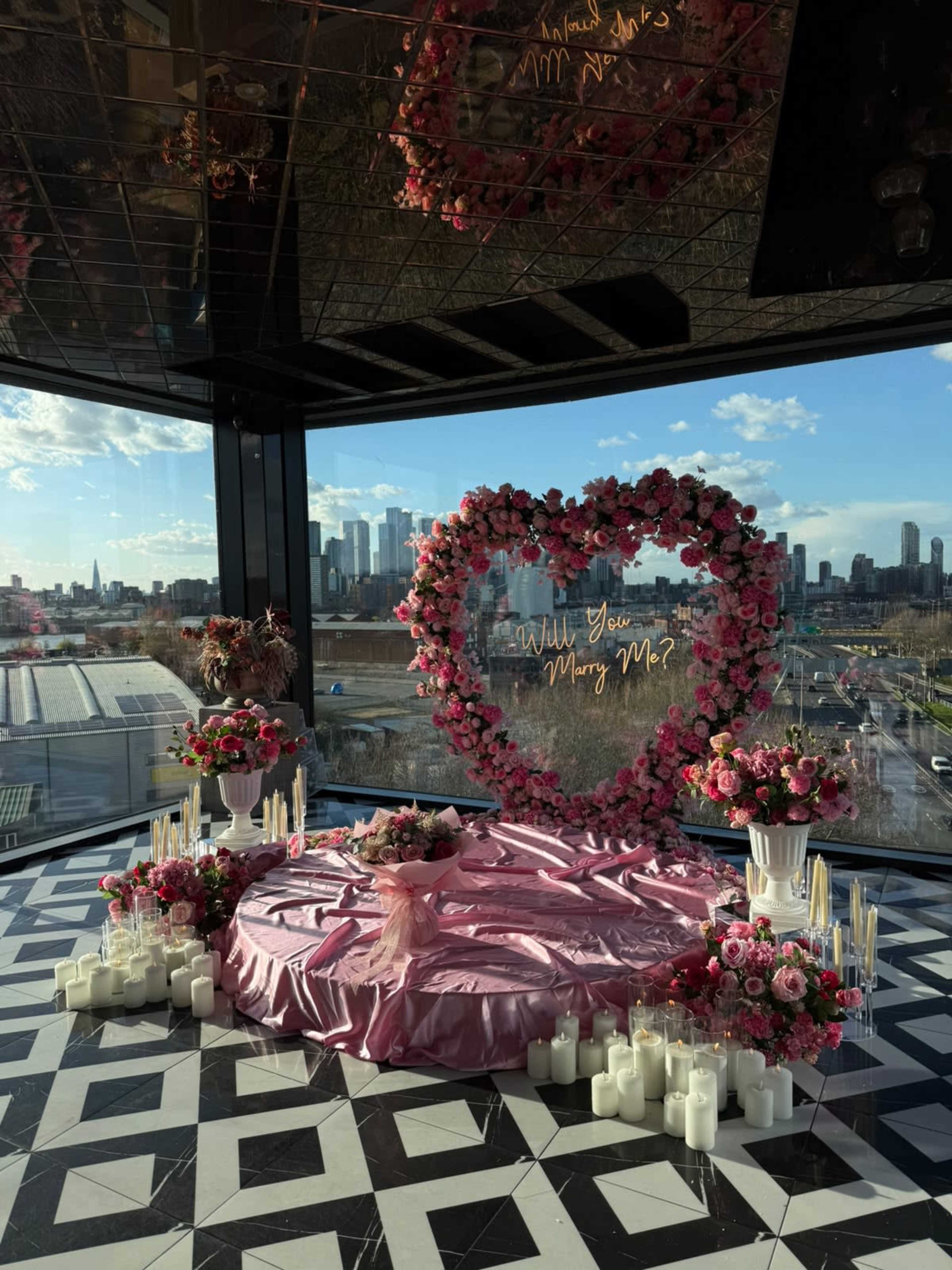 The image depicts a romantic proposal setting featuring a heart-shaped floral arrangement, surrounded by candles and a pink tablecloth, with a city skyline visible in the background.