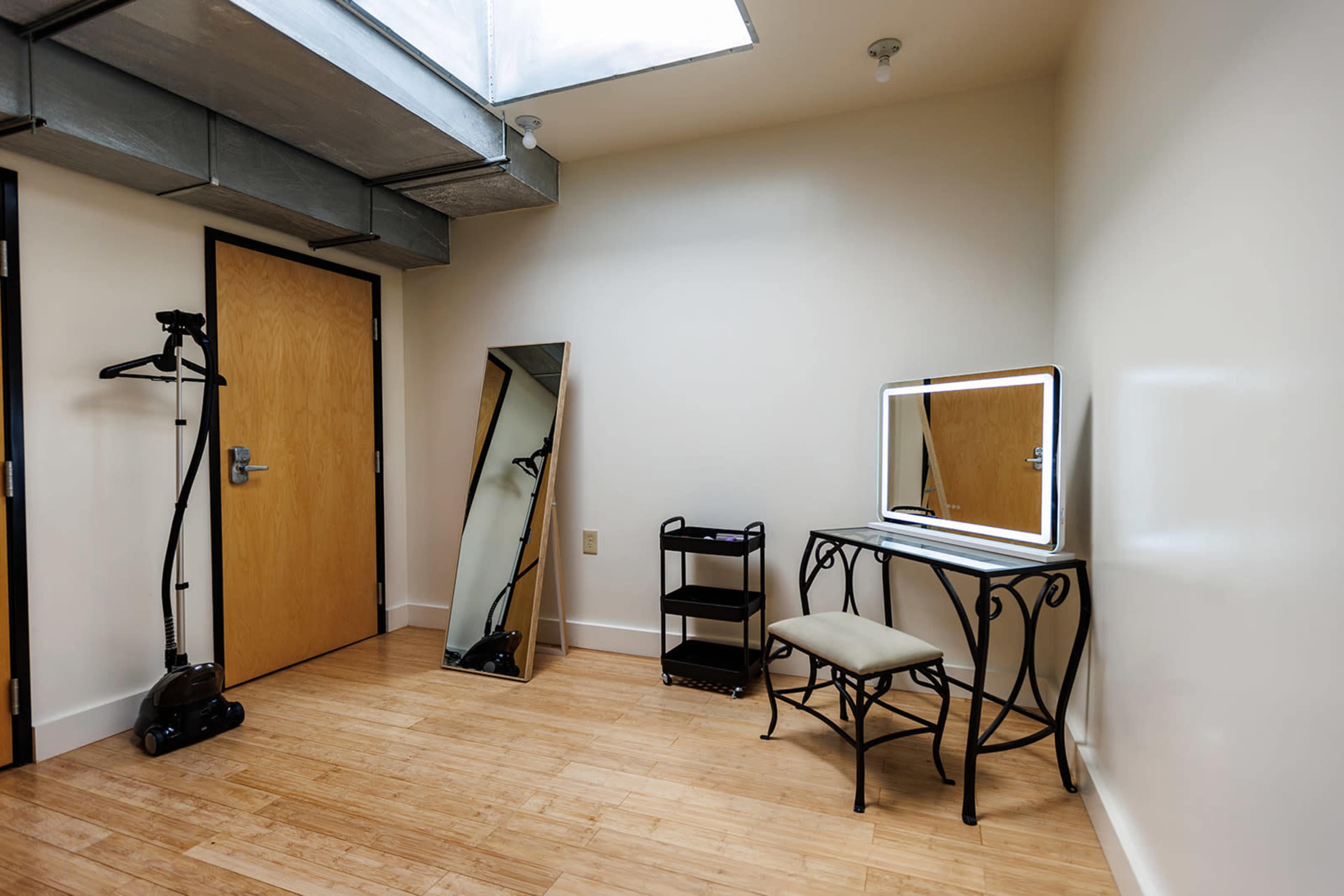 The image shows a minimalist room featuring a wall-mounted mirror, a small chair, a vanity table with lights, a clothing rack, and a black shelving unit.