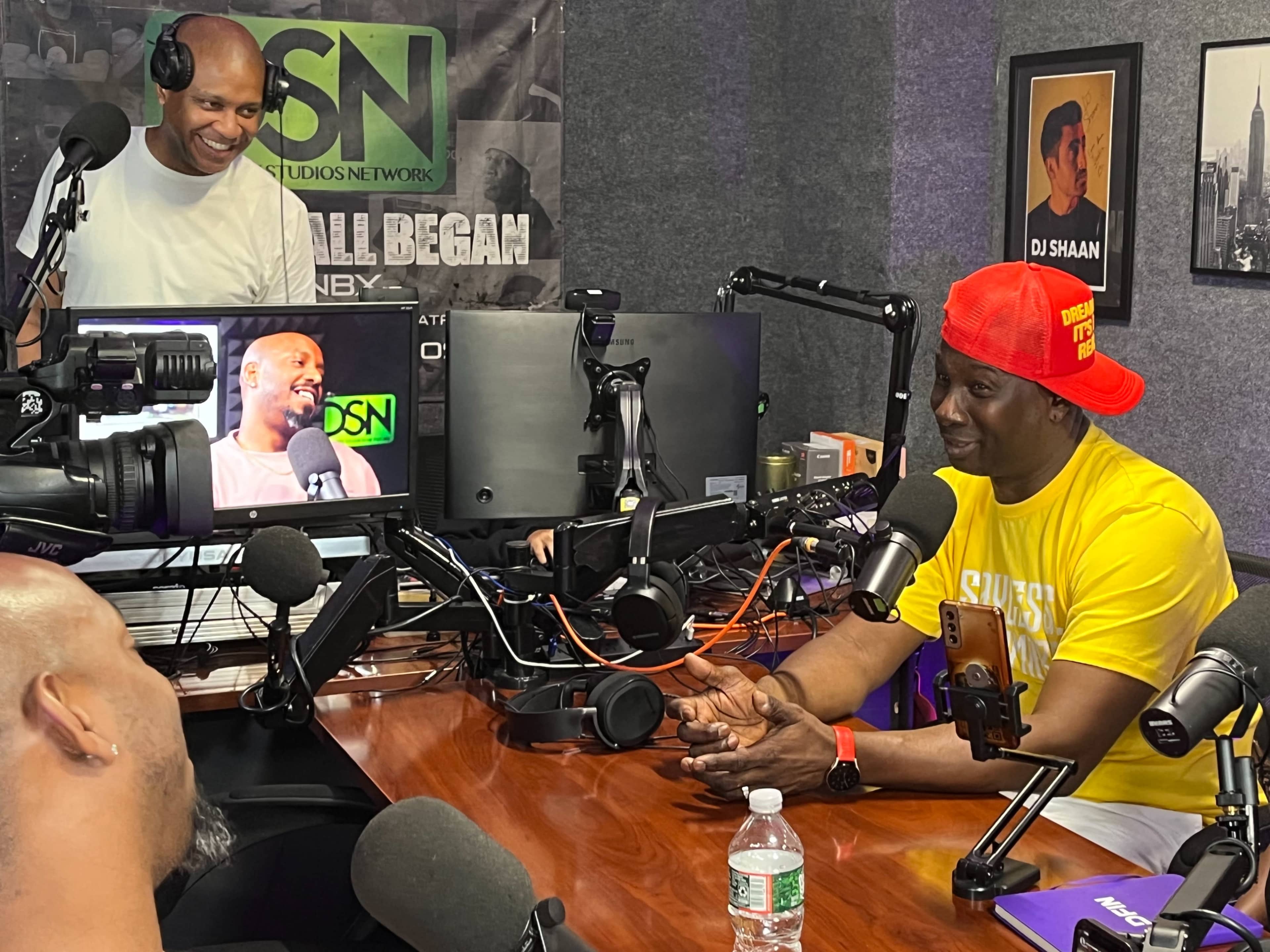 Two individuals are seated at a podcast studio, one speaking while the other operates a camera and monitors the recording.