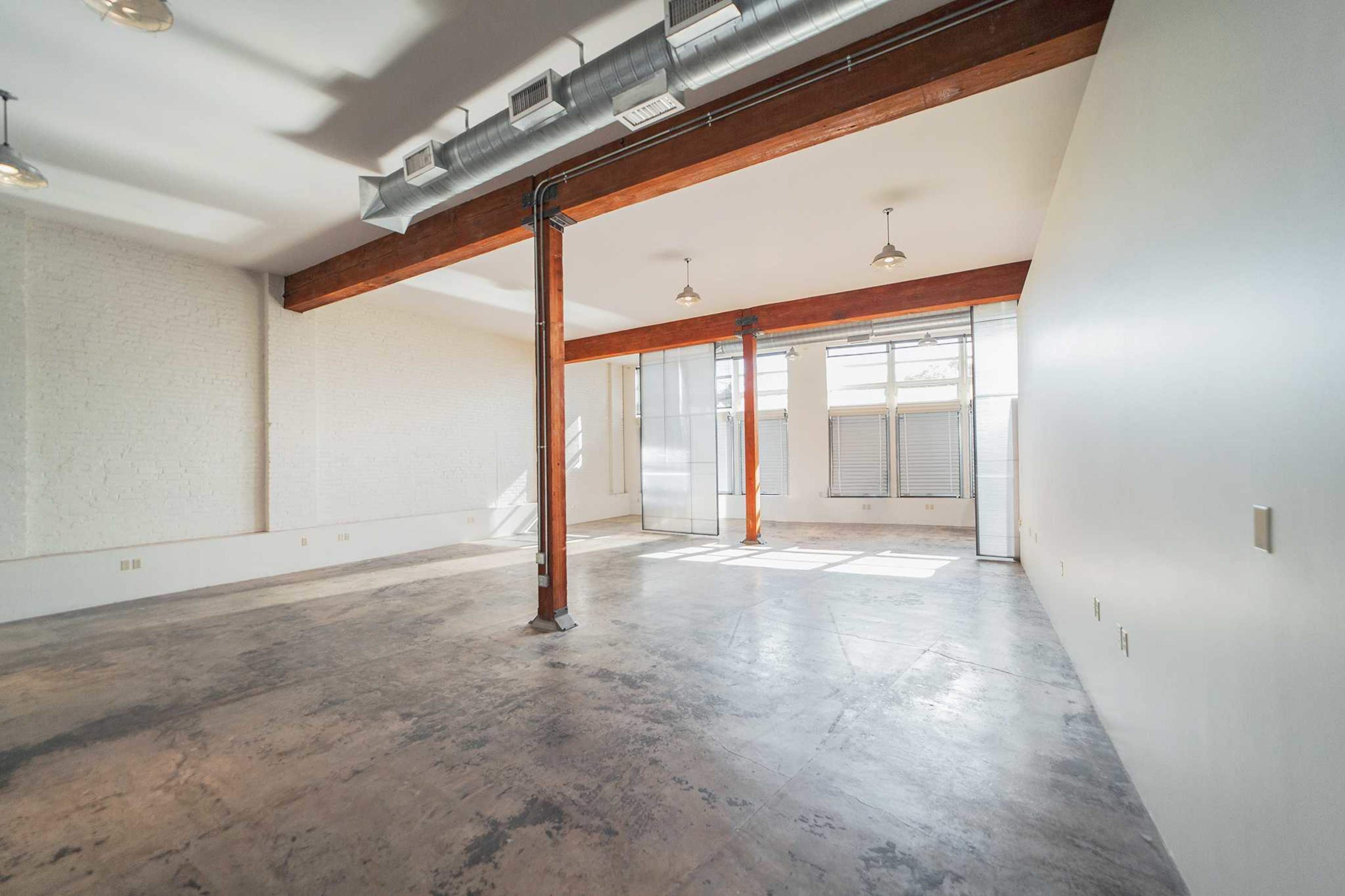 The image shows a spacious, empty room with large windows, exposed wooden beams, and concrete flooring.