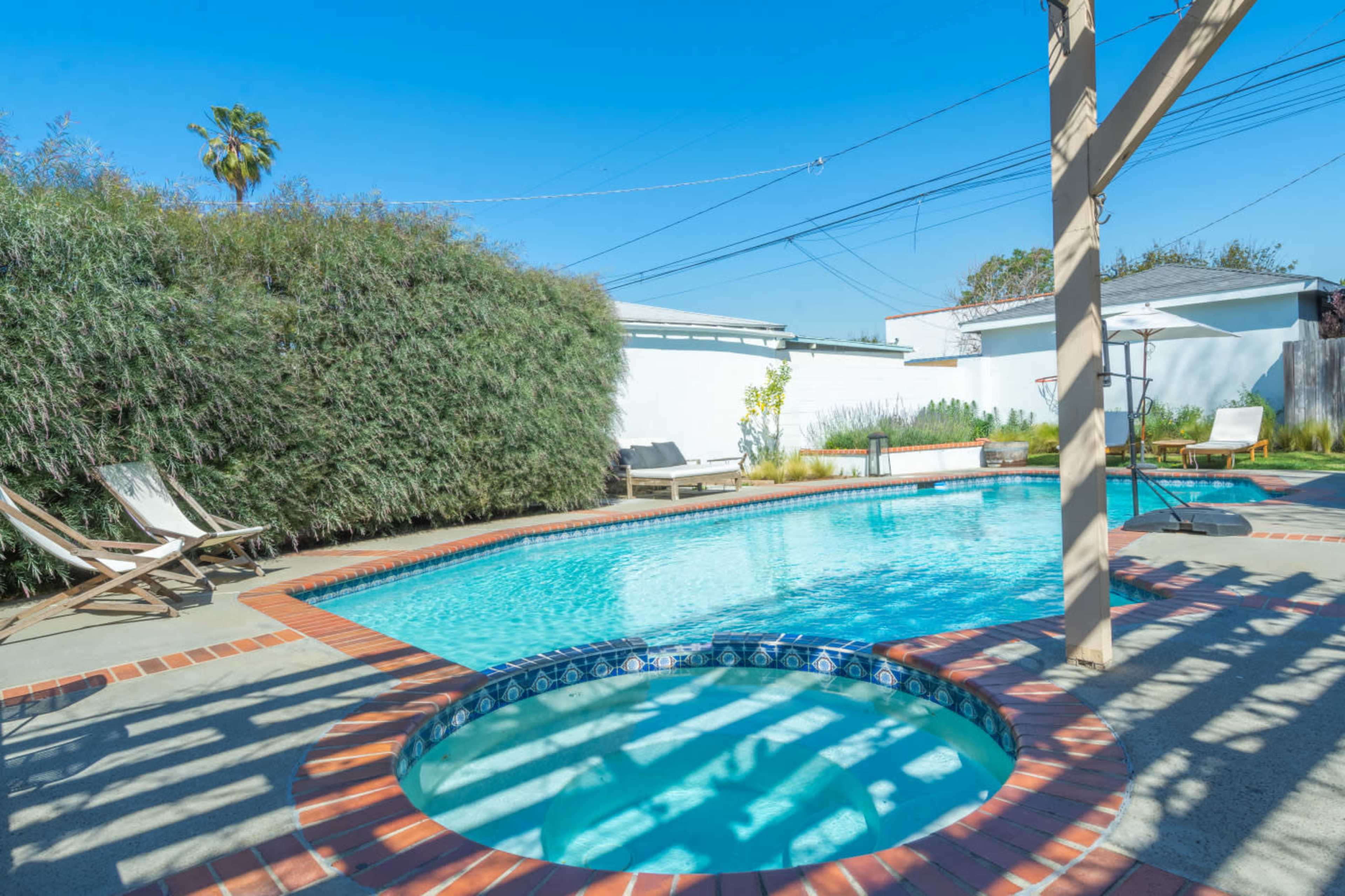 A clear swimming pool with a spa is surrounded by seating areas and greenery under a blue sky.