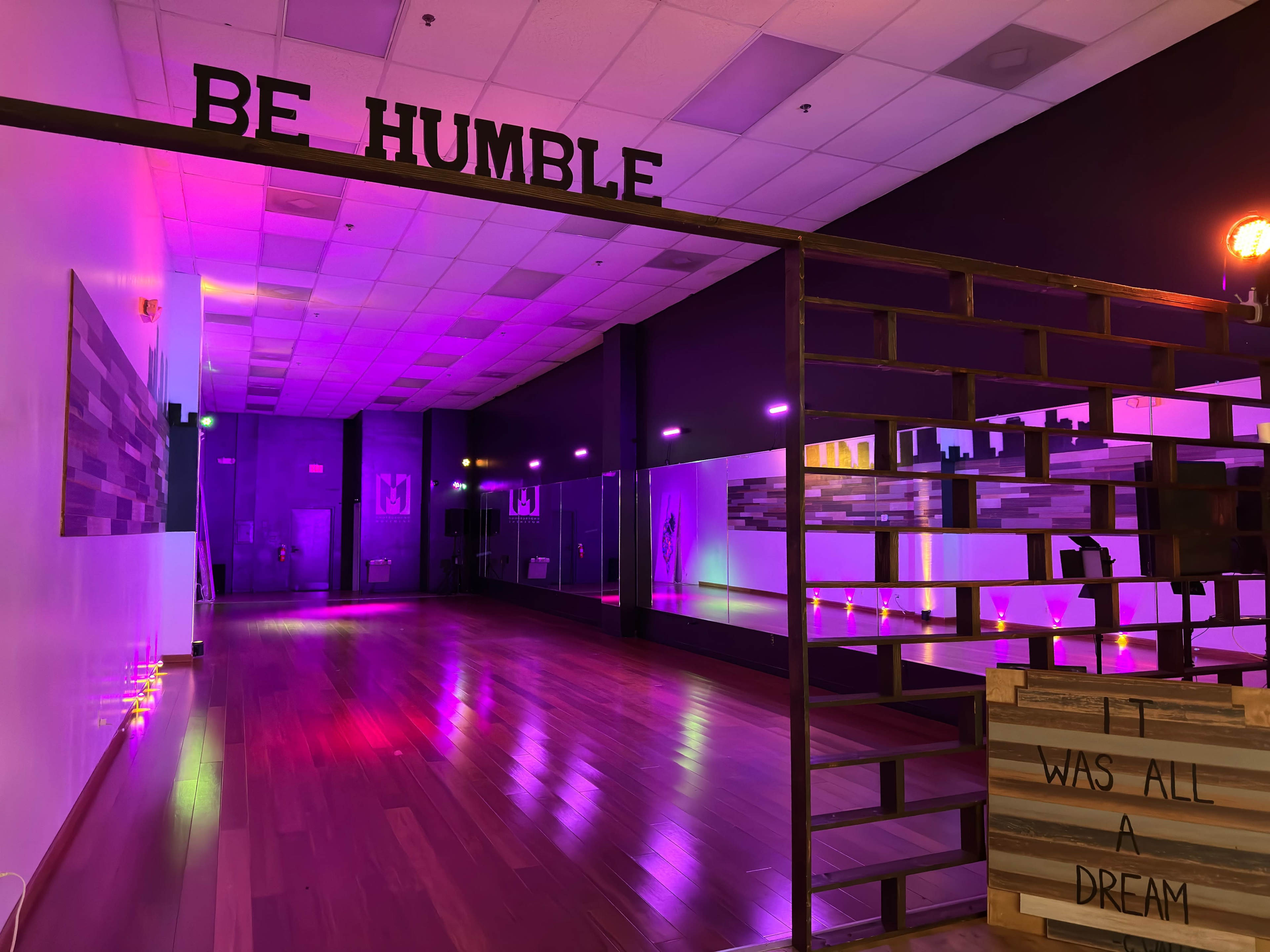 A brightly lit dance studio with colorful lights and a sign that reads "BE HUMBLE" near an entrance.