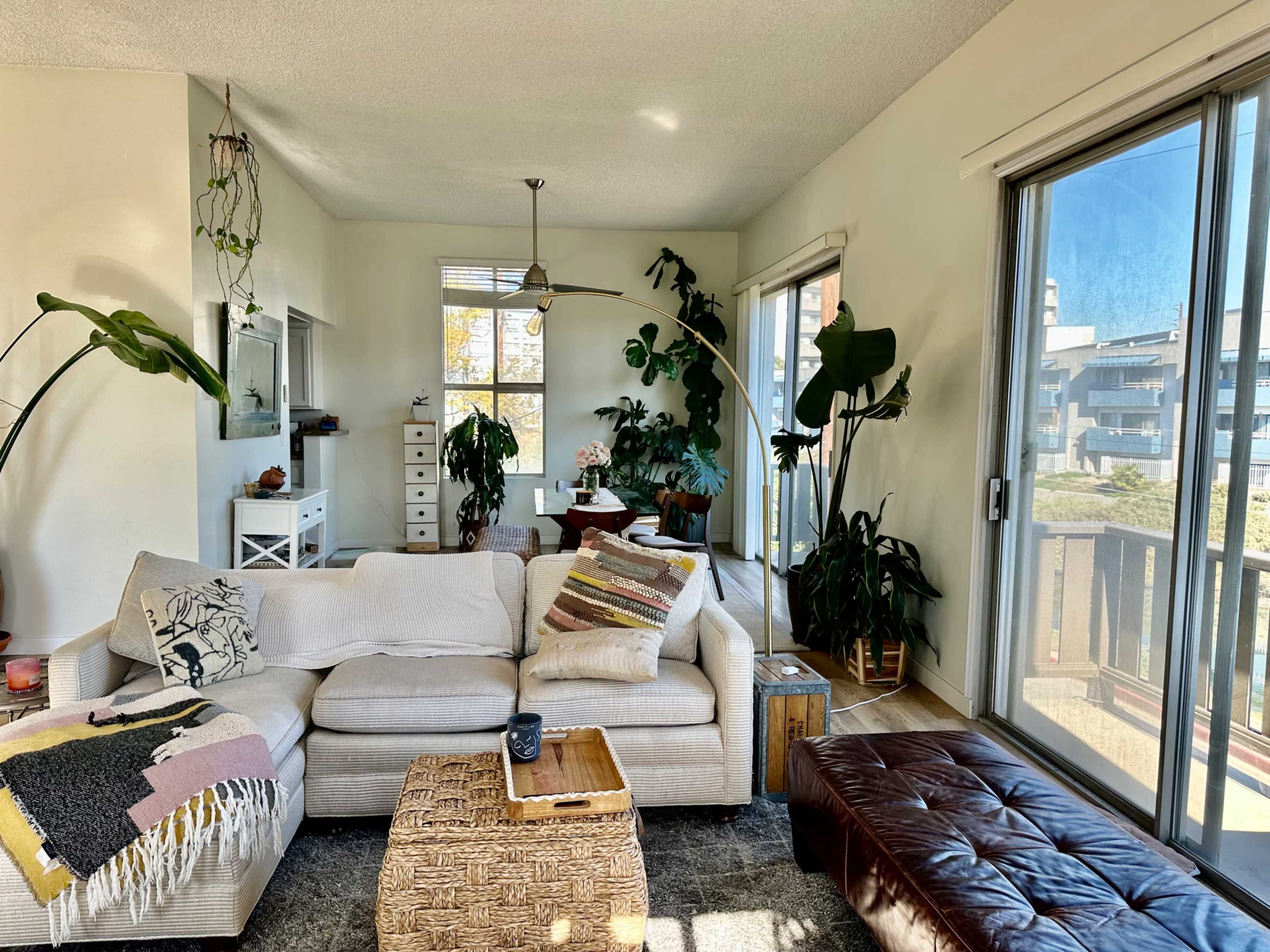 The living room features a light-colored sectional sofa adorned with decorative pillows, a woven coffee table, and large plants near floor-to-ceiling windows.