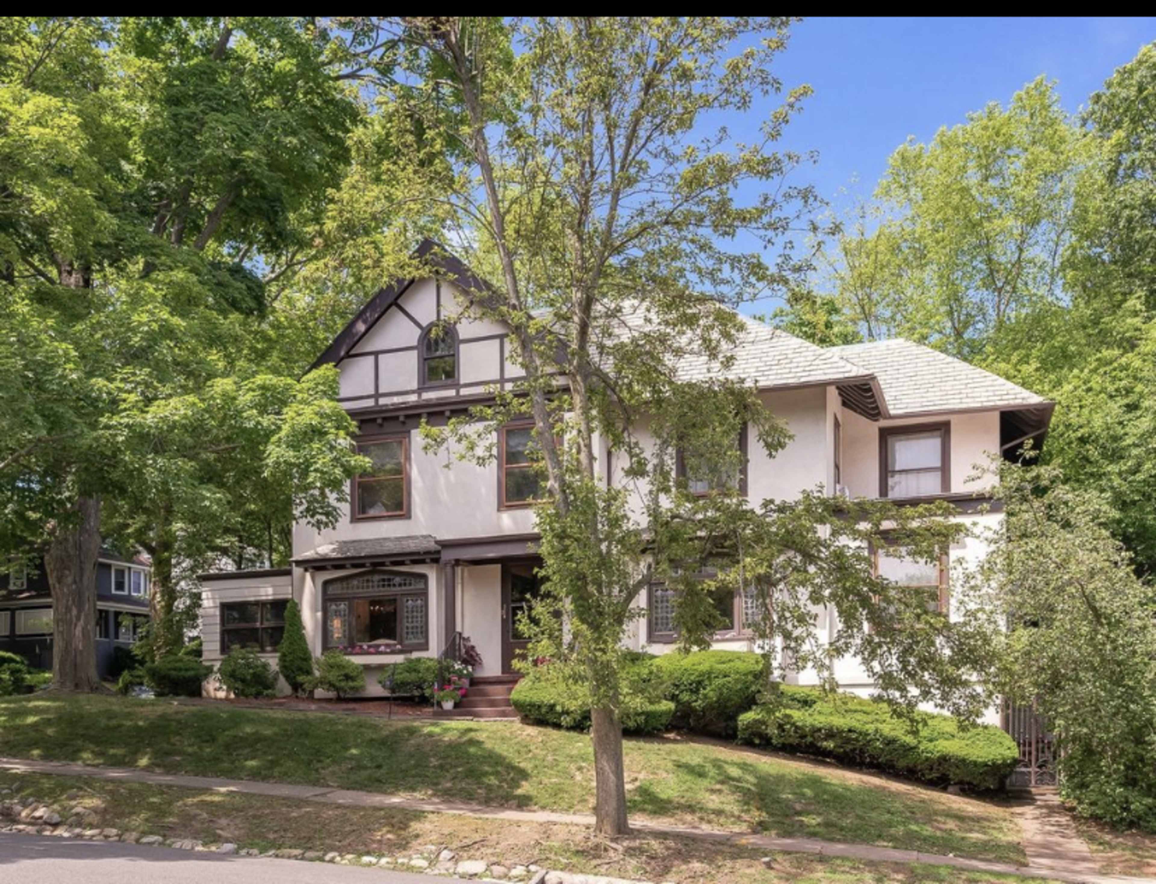 The image shows a large, multi-story house with a combination of white and dark brown siding, surrounded by green trees and shrubs.