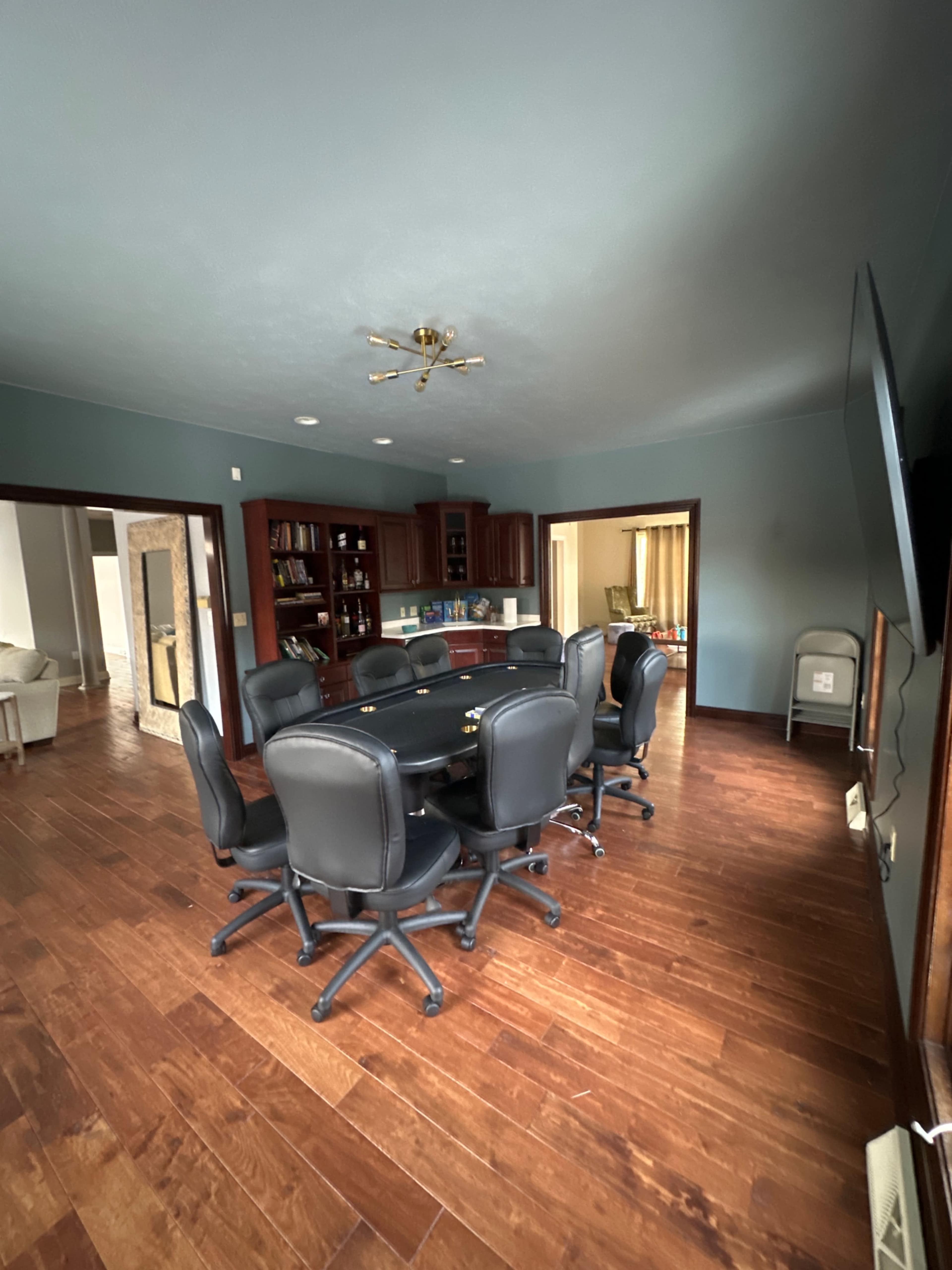 A large meeting room features a round black poker table surrounded by eight black swivel chairs, with a kitchen area visible in the background.