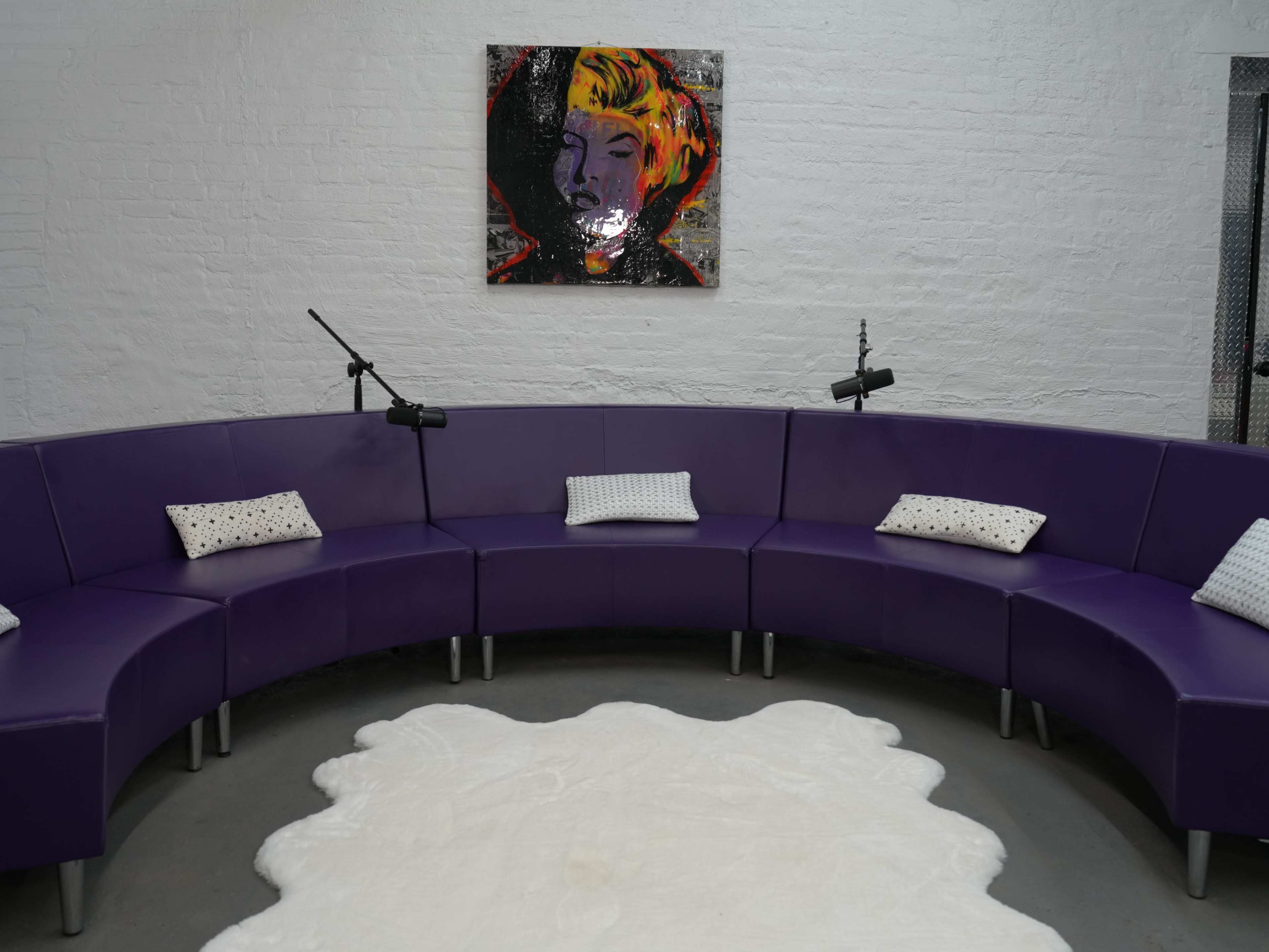 The image shows a curved purple seating arrangement with white pillows and a round white rug, accented by a colorful artwork on the wall.
