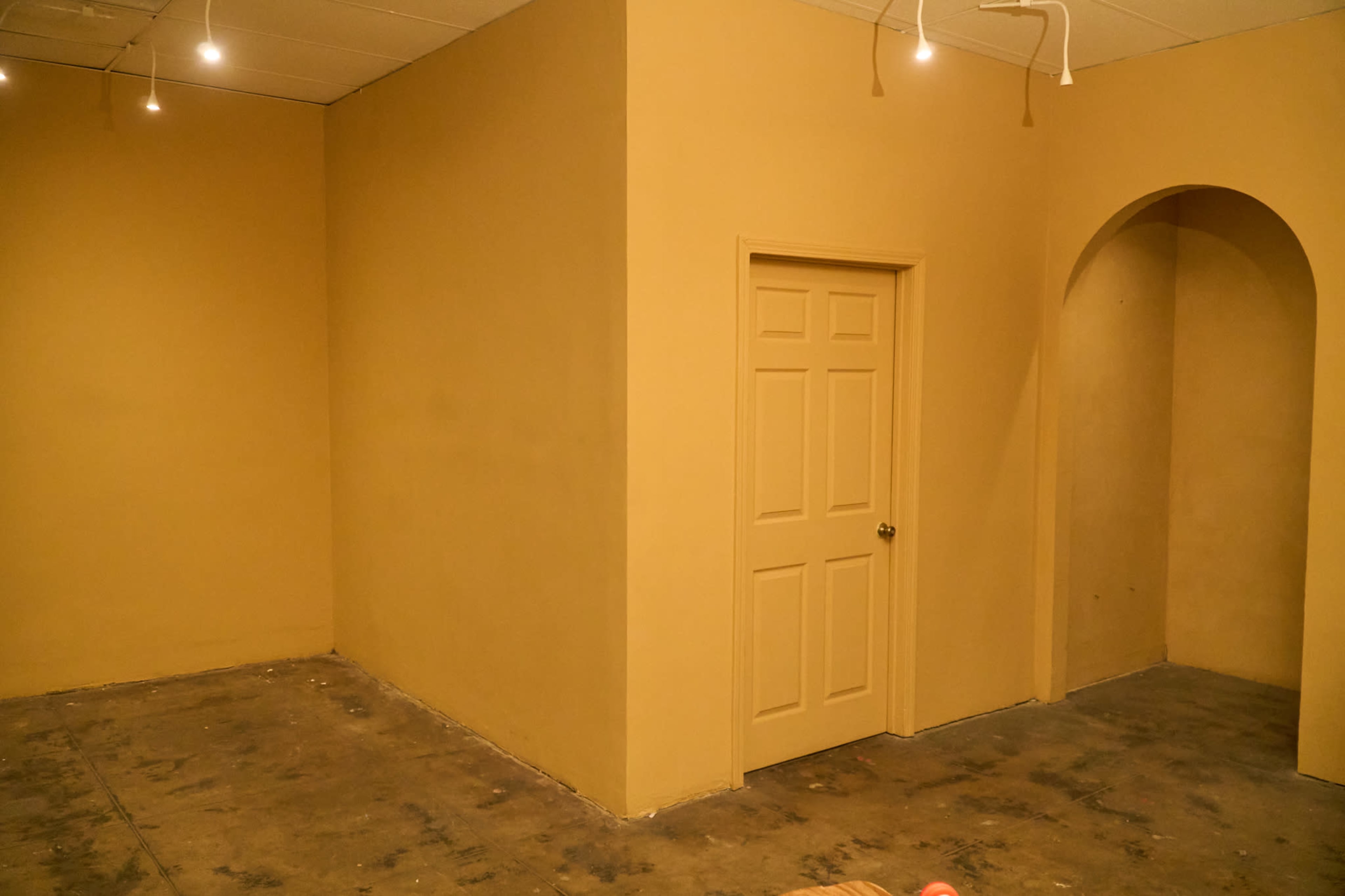 The image shows an empty room with beige walls, a closed door in the center, and an arched opening on the right side.