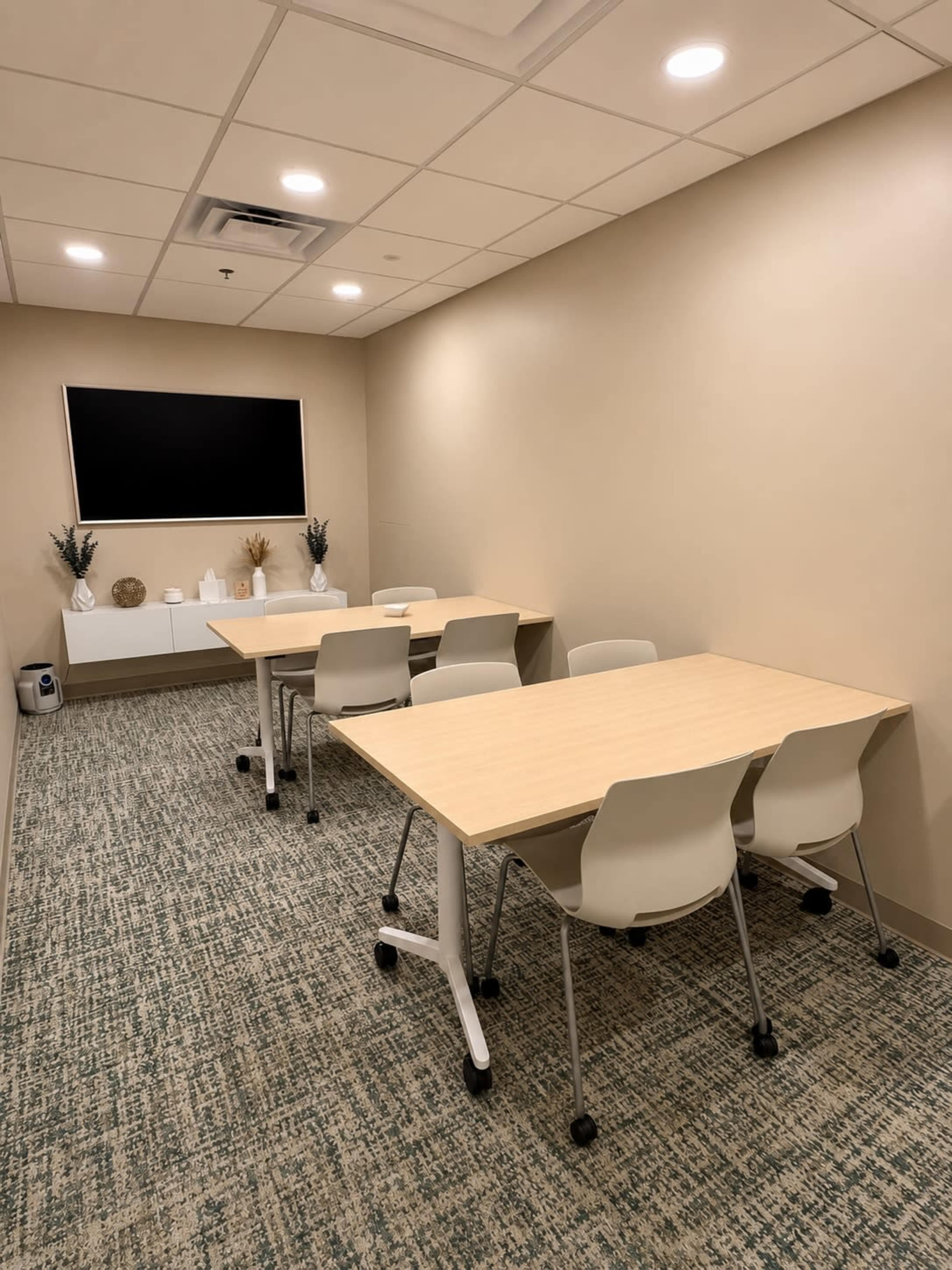 The image shows a light-colored conference room with two tables arranged for meetings, four chairs around each table, and a wall-mounted screen.