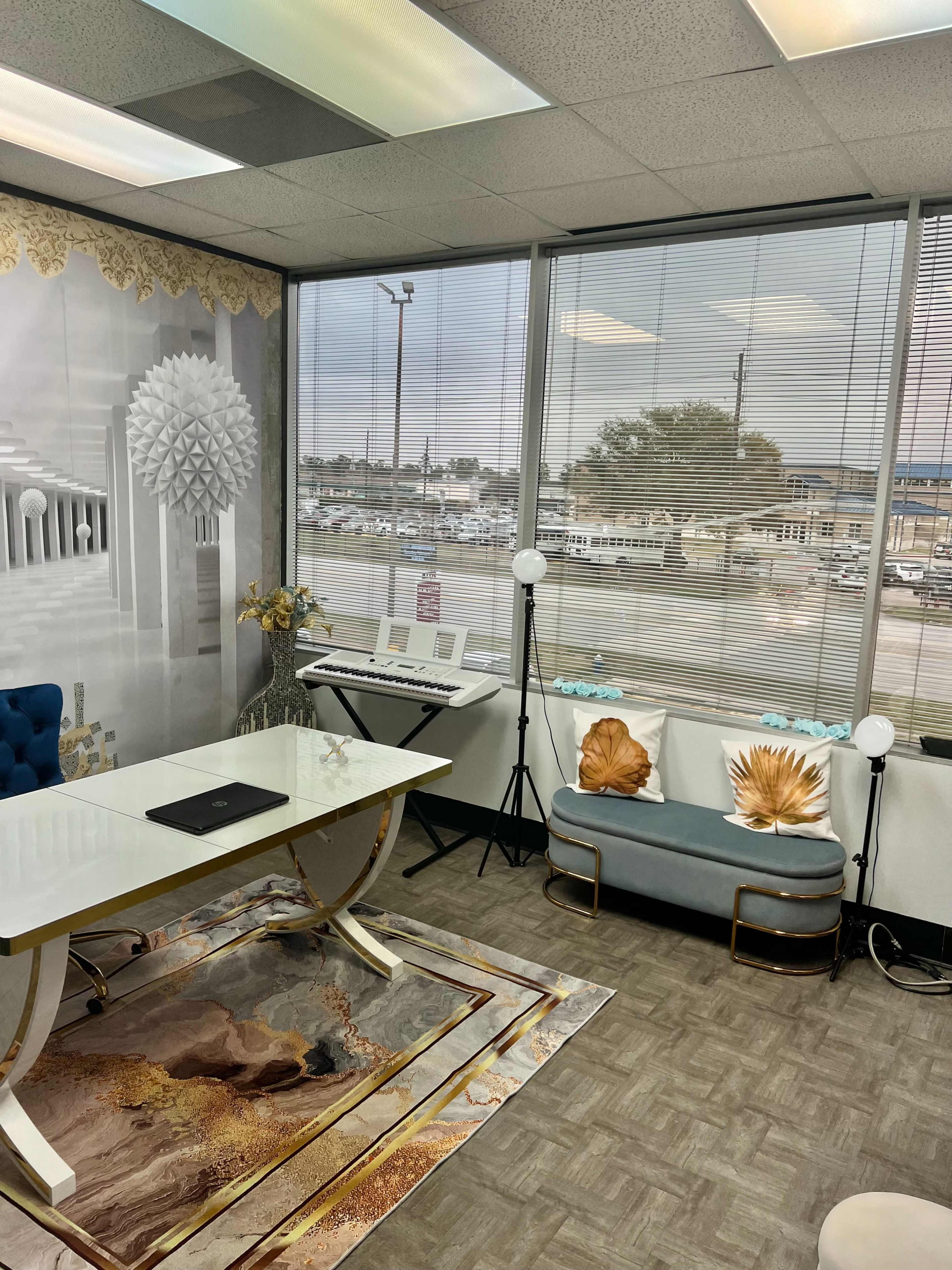 The image shows a modern office space with a white table, blue seating, decorative elements, and a view of an outside parking lot through large windows.