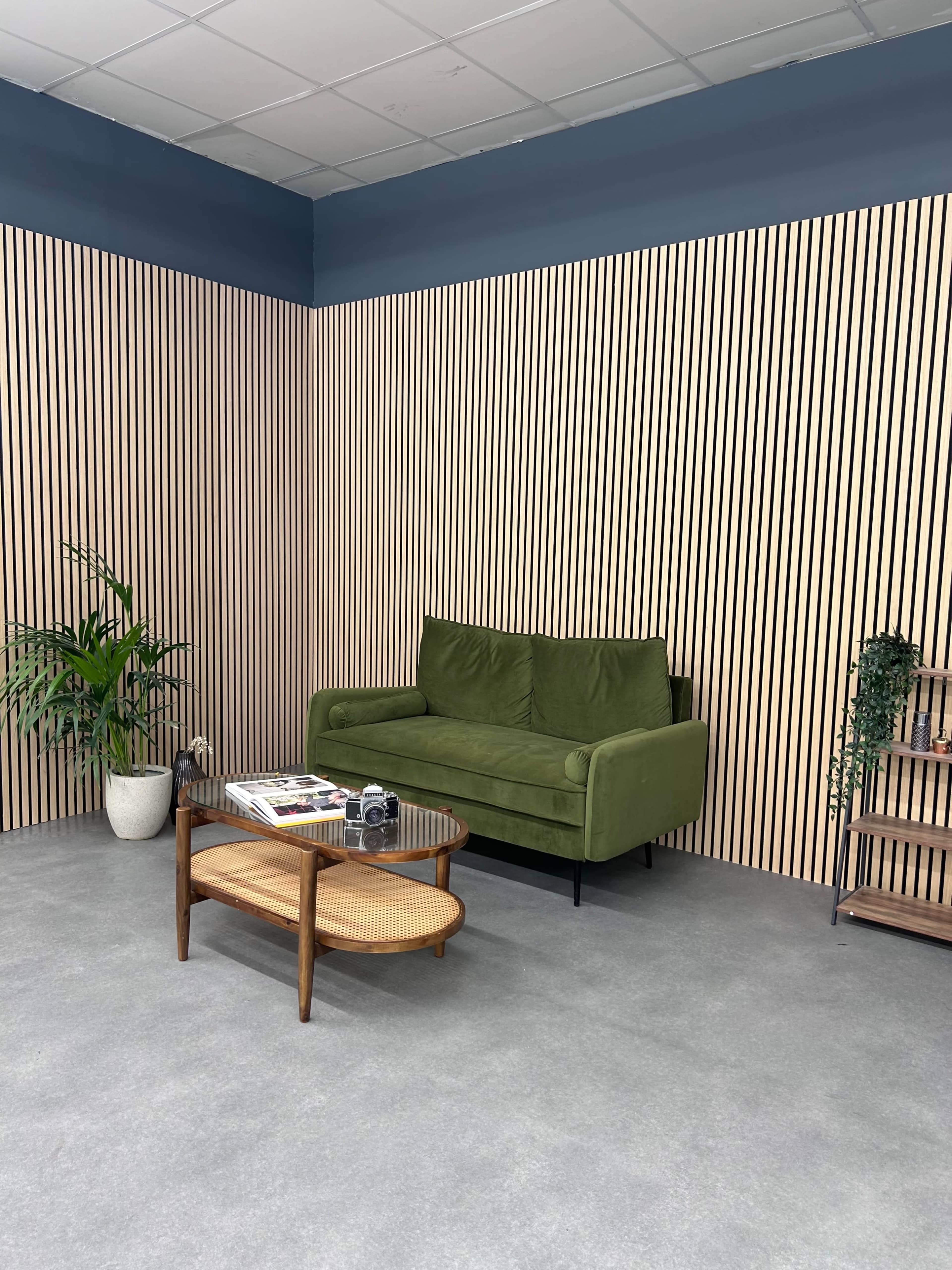 A modern lounge area featuring a green sofa, a coffee table, a plant, and a wooden shelf against a striped wall.