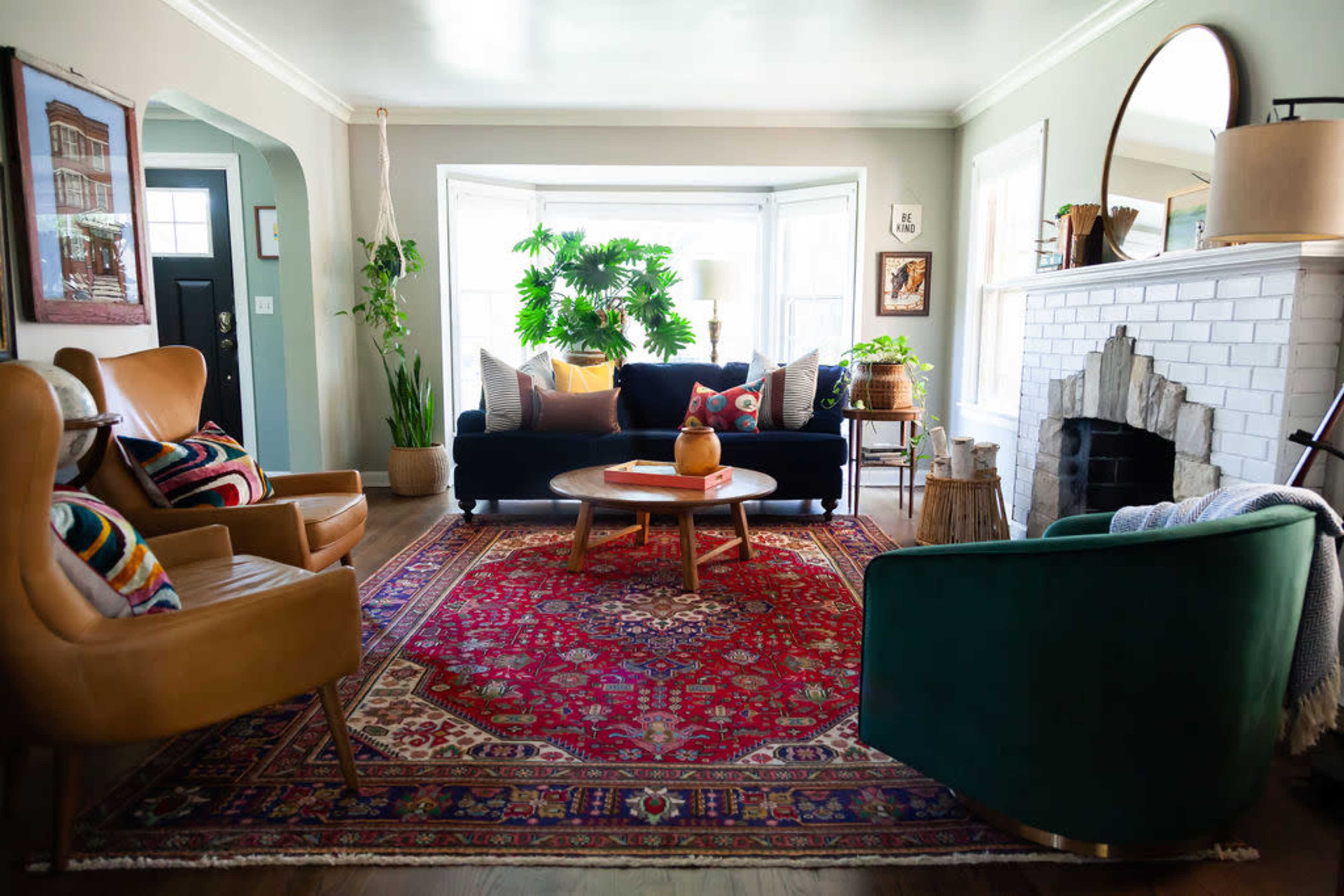 A cozy living room features a dark sofa and colorful armchairs set around a wooden coffee table on a patterned rug, with large windows allowing natural light to fill the space.