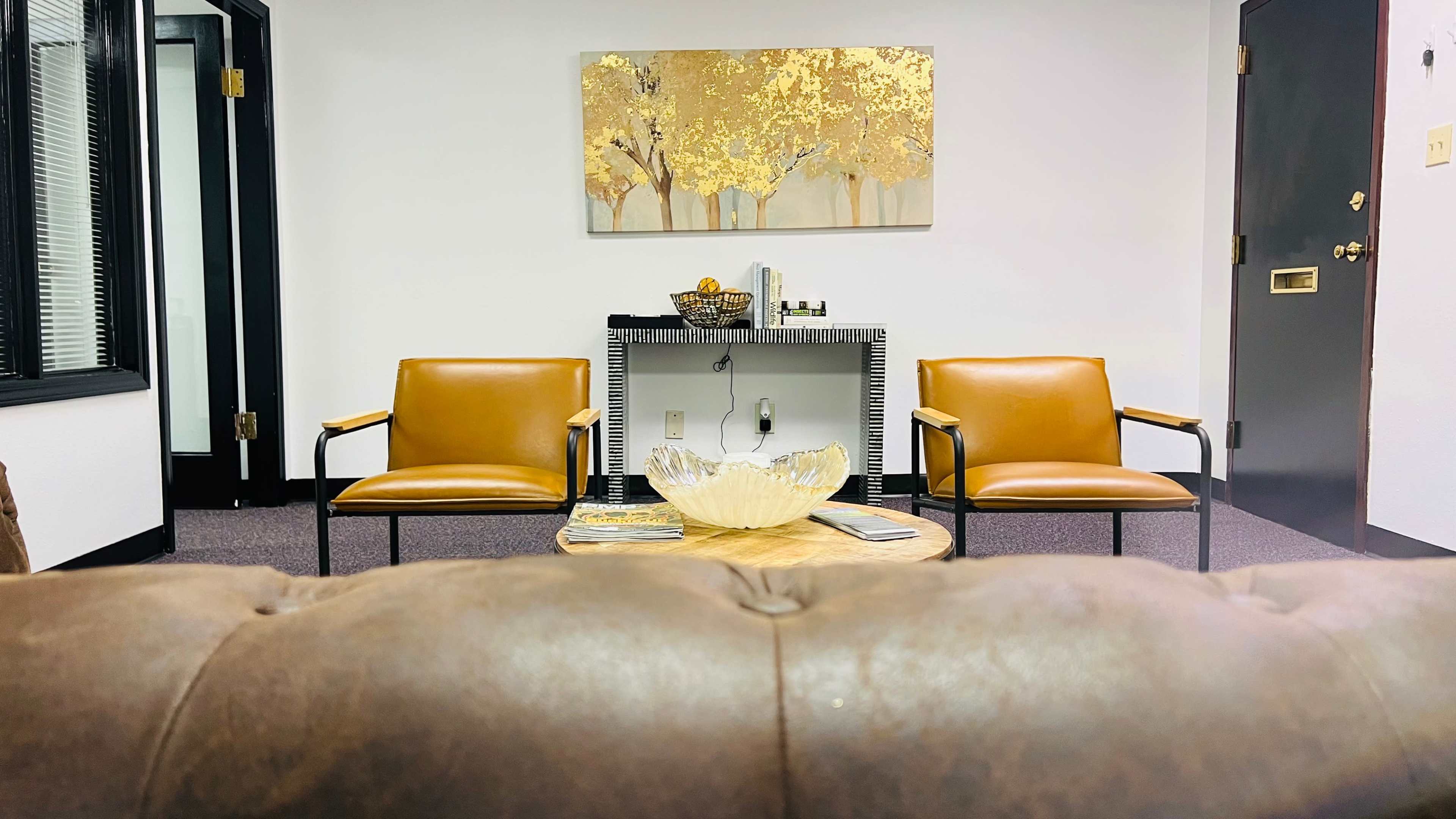 A modern office waiting area with two brown chairs facing a small table, adorned by a decorative bowl and framed art on the wall.