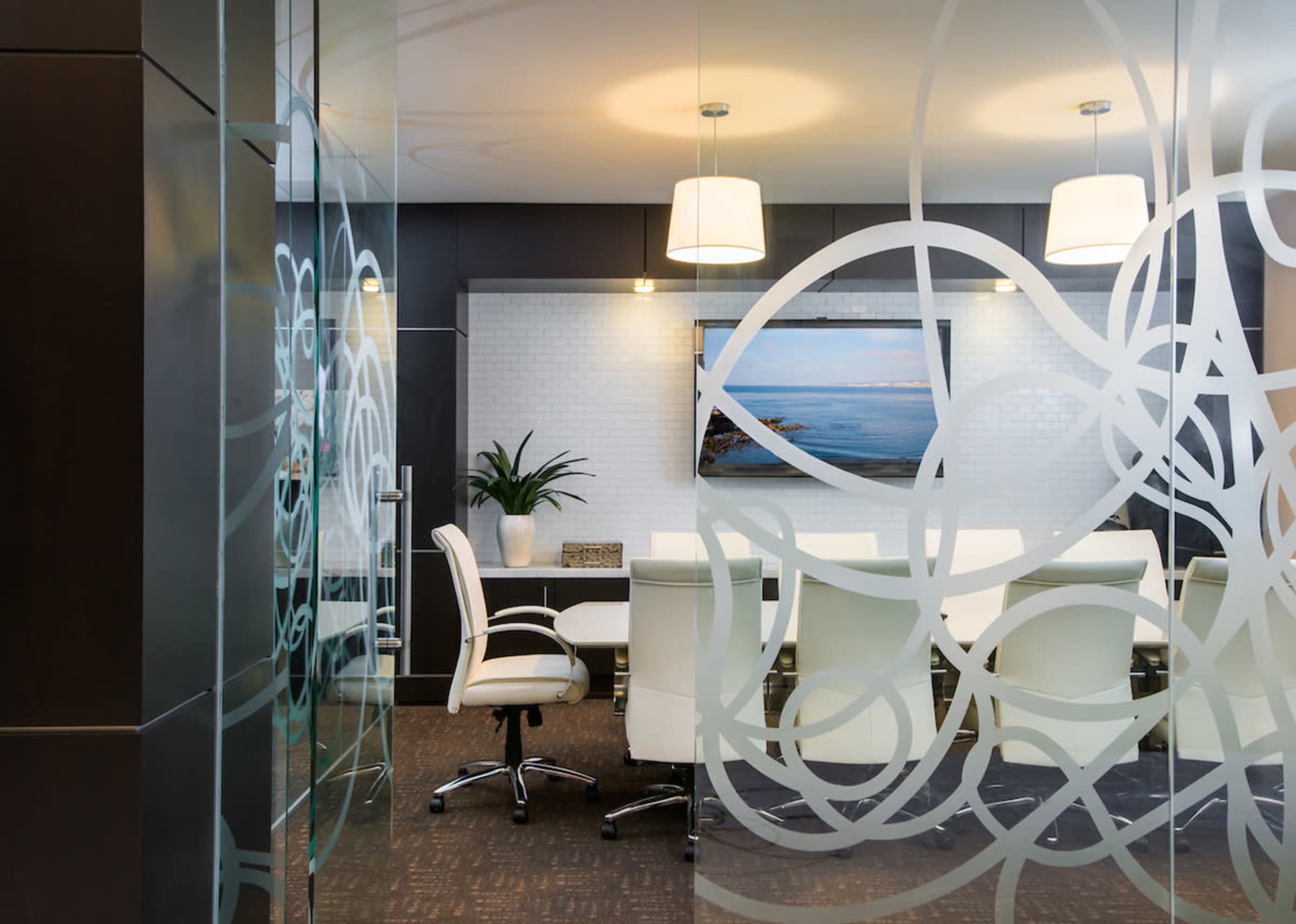 A conference room features a large white table surrounded by chairs, with a television displaying a coastal image and decorative lighting overhead.