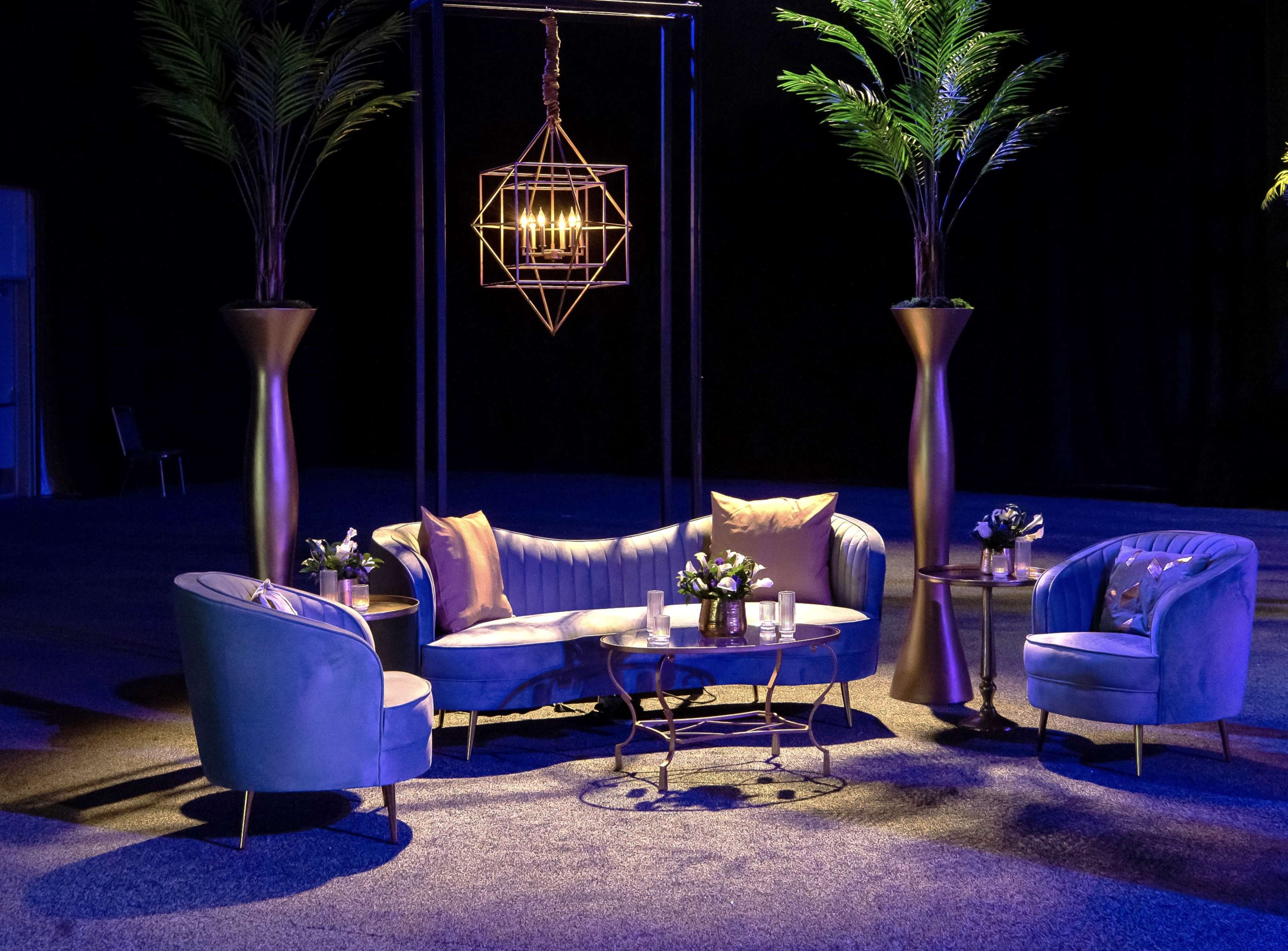 The image shows a stylish lounge area with blue velvet furniture, gold accents, and potted plants under soft lighting.