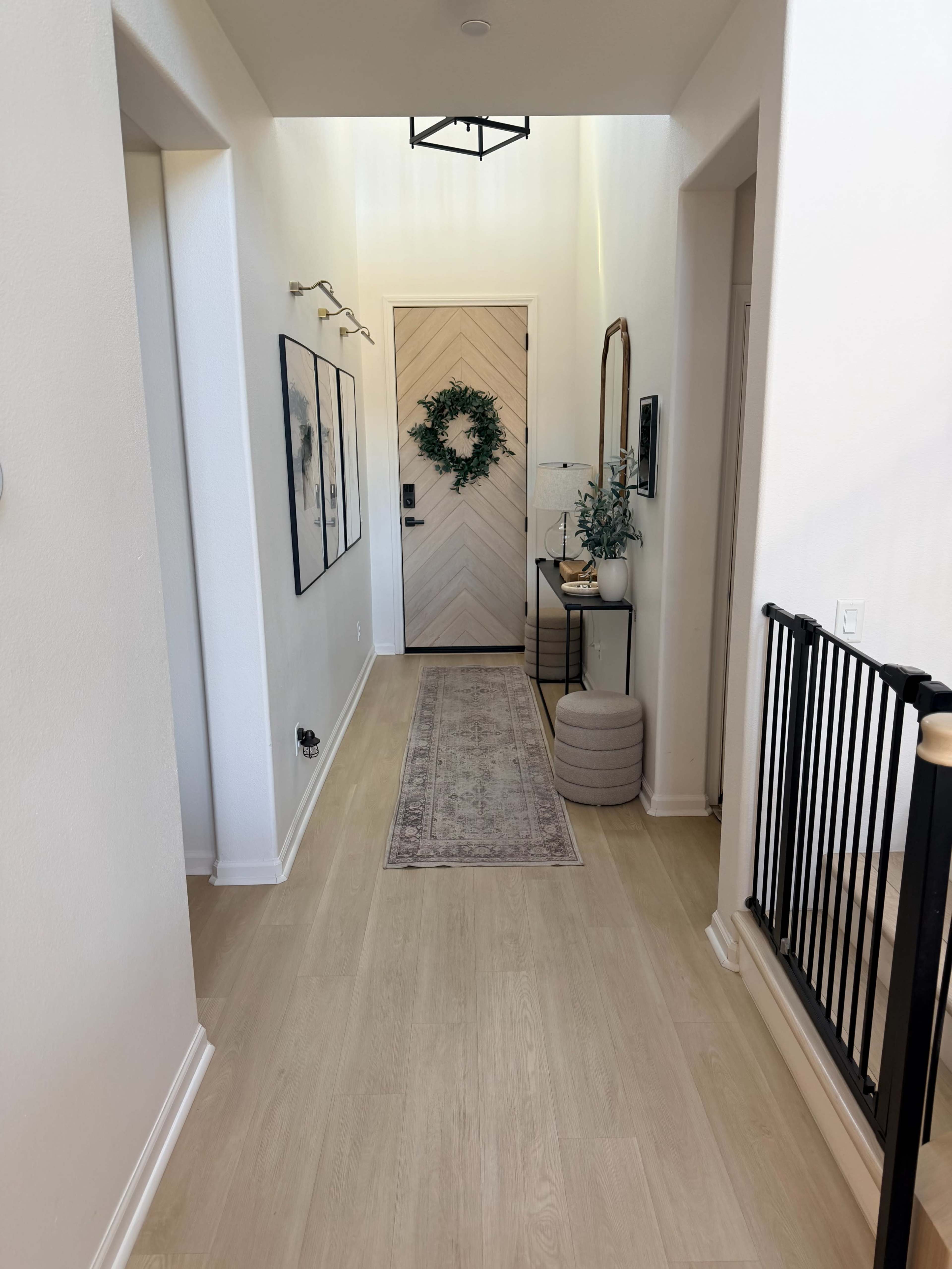The image shows a well-lit hallway with light wood flooring, a front door at the end, and a console table adorned with a plant and mirror along one side.