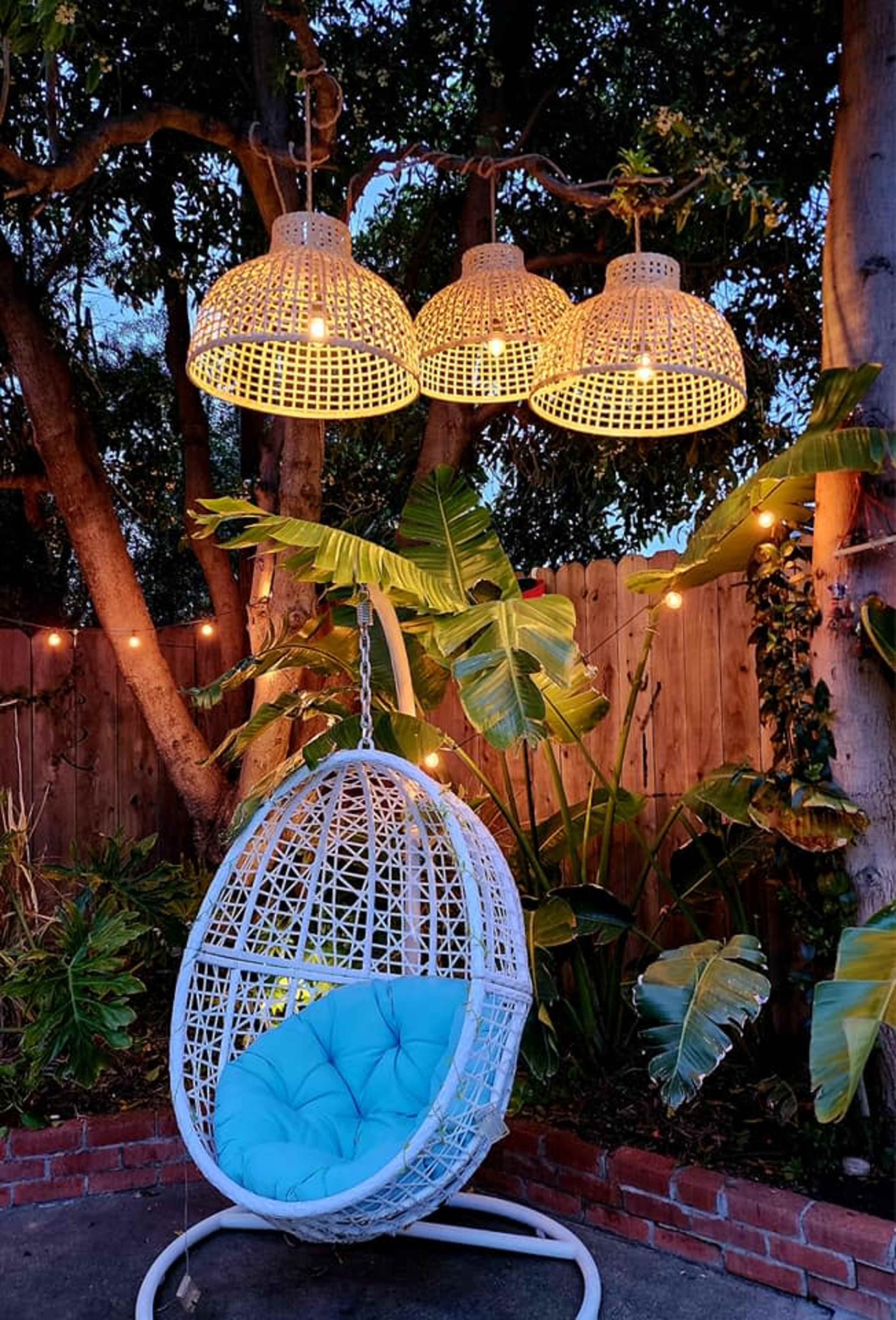 A white hanging chair with a blue cushion is positioned in a garden under three woven light fixtures and surrounded by tropical plants.