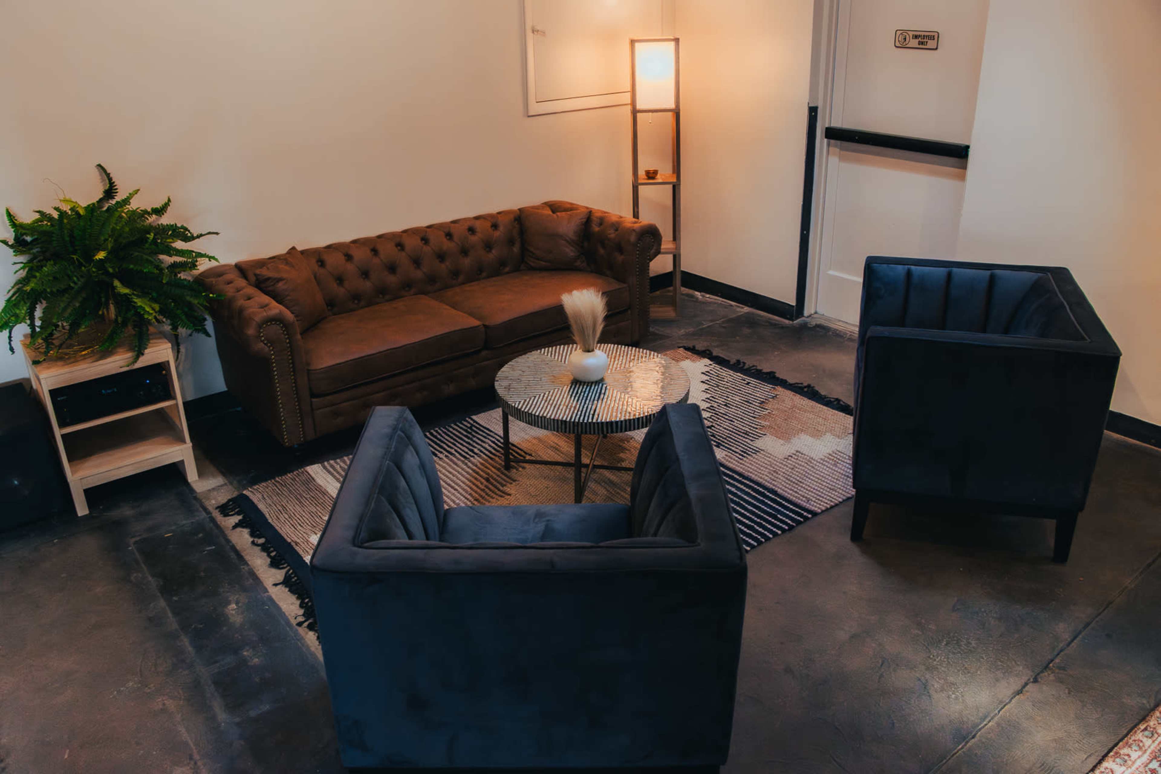 The image shows a modern lounge area with a brown leather couch, two blue velvet chairs, a round glass coffee table, and a potted plant.