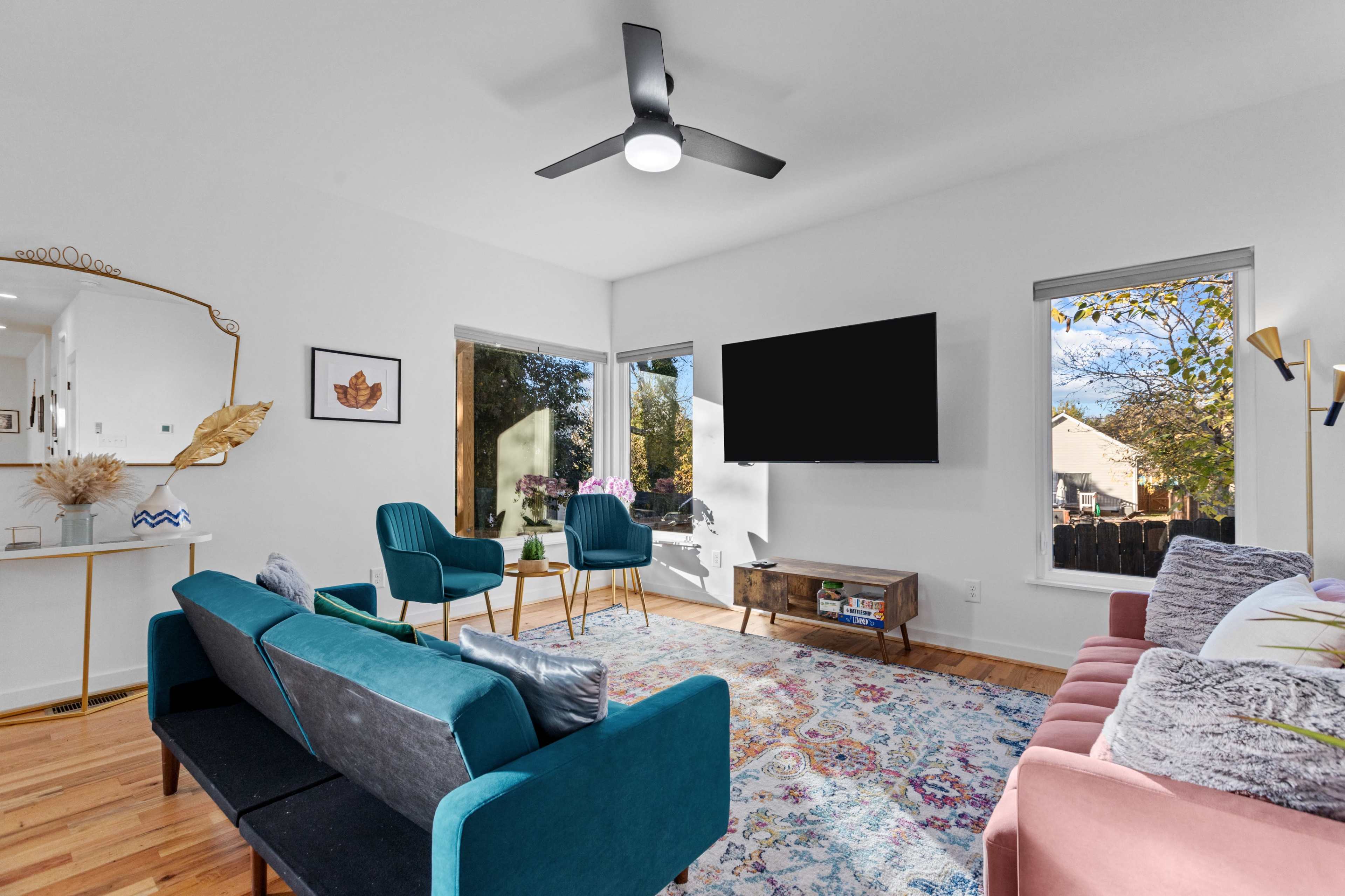 A modern living room features a mix of teal and pink sofas, a wooden coffee table, and a wall-mounted television, with large windows allowing natural light to fill the space.