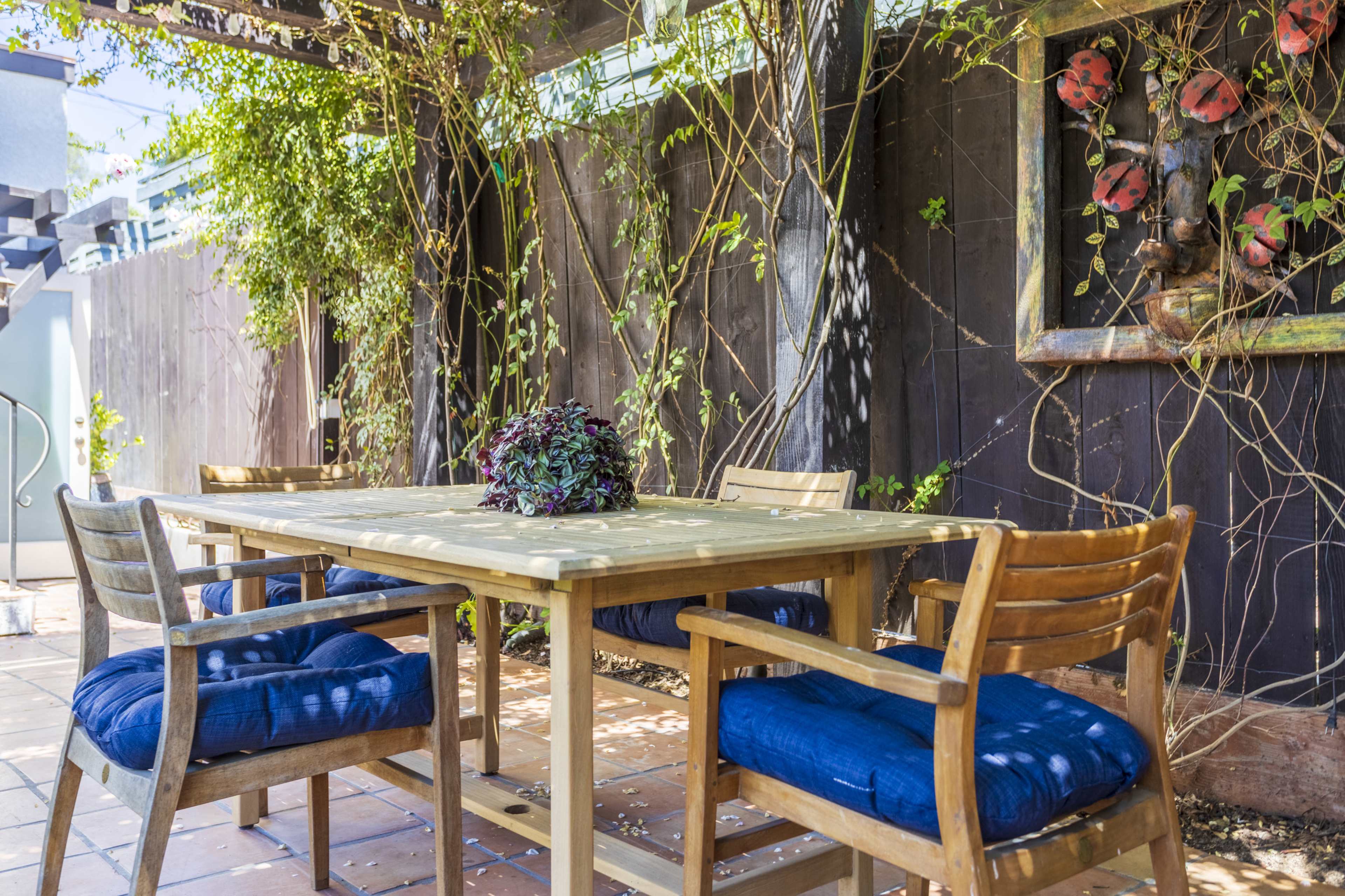 A wooden dining table with blue cushions on the chairs is set under a trellis adorned with vines and flowers in an outdoor patio area.