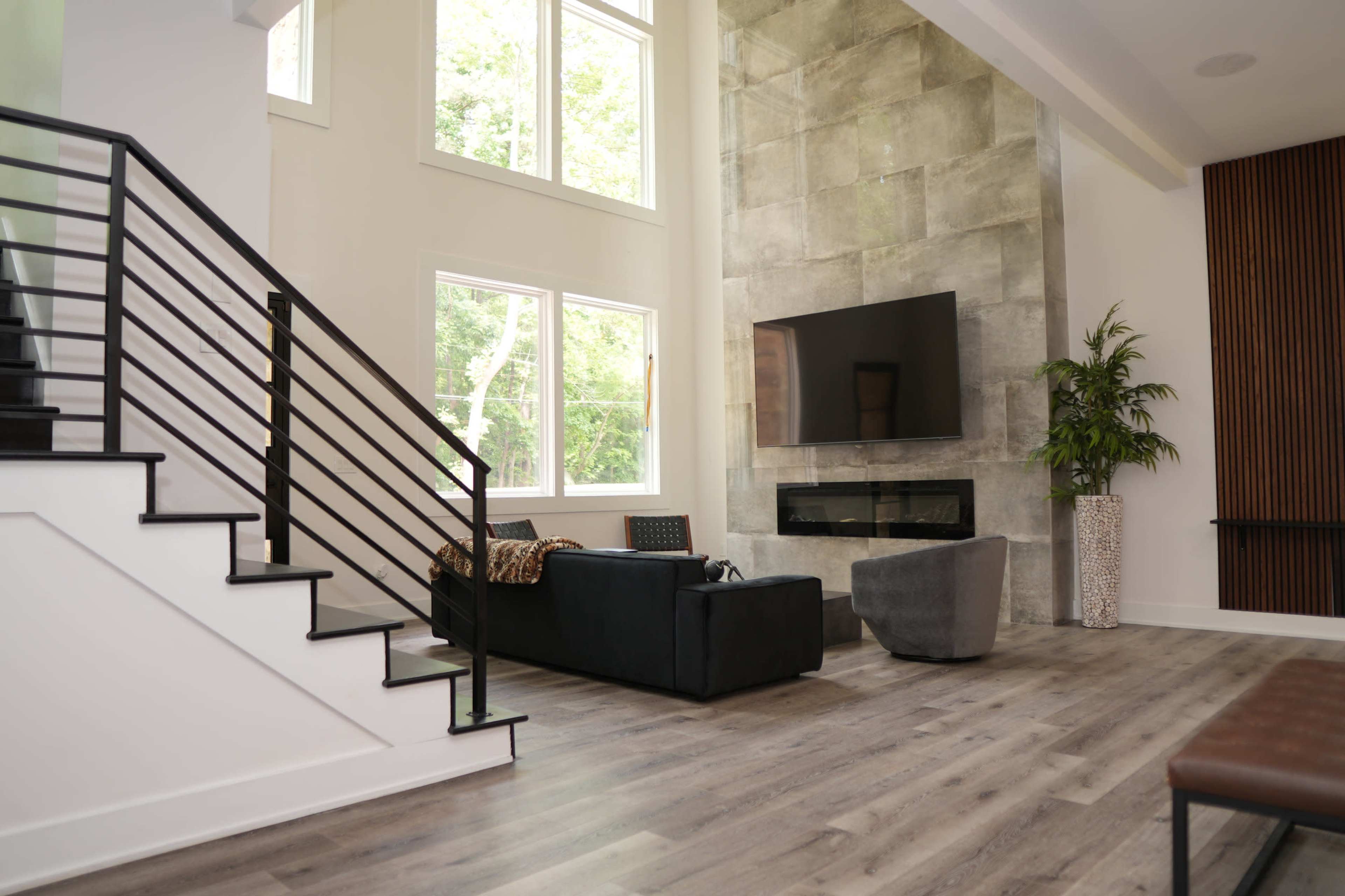A modern living room with large windows, a stone accent wall, a mounted television, and a staircase leading to the upper floor.