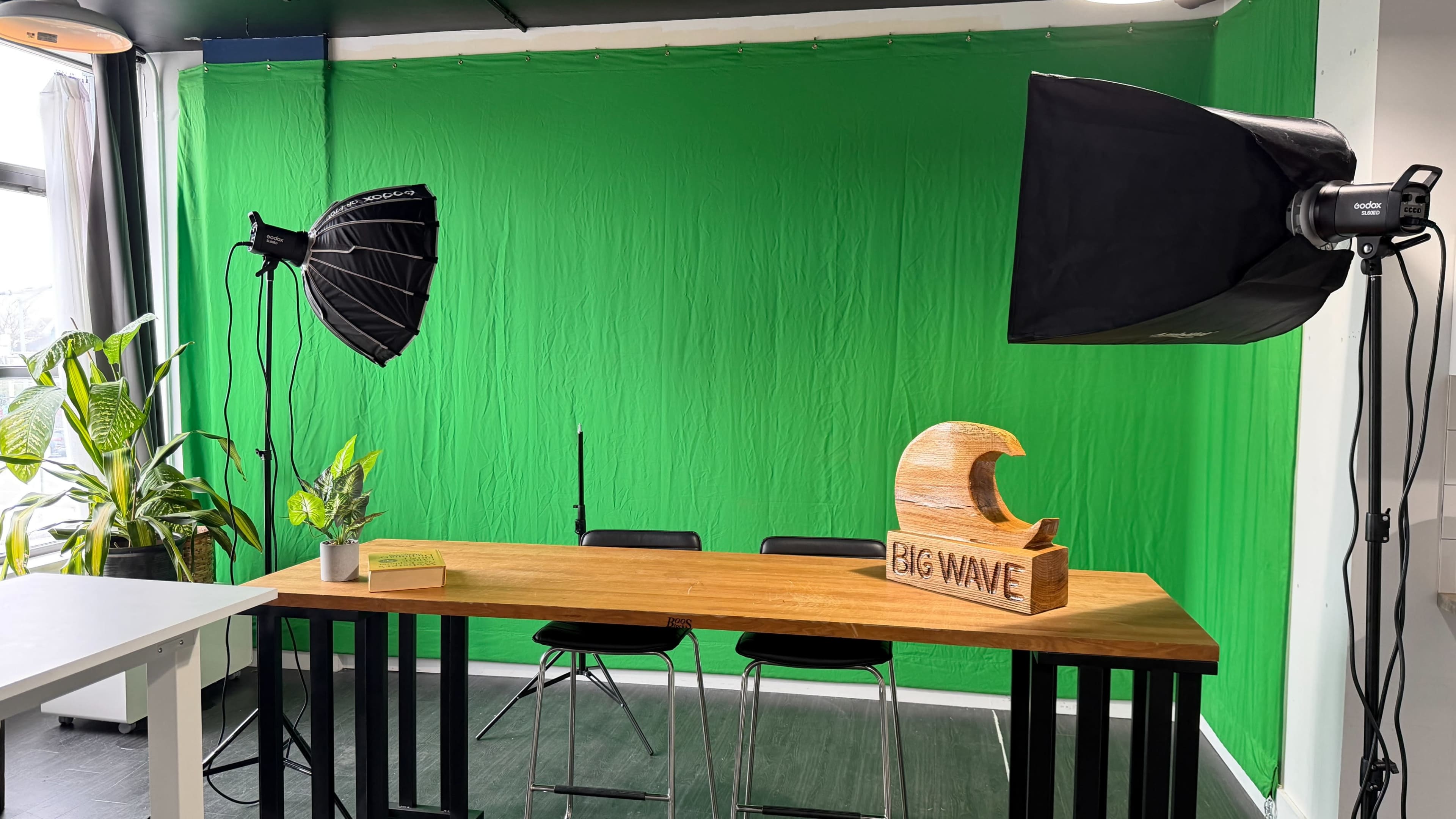 The image shows a studio setup with a green screen backdrop, a wooden table with a decorative wave sculpture labeled "BIG WAVE," two softbox lights, and two black chairs.