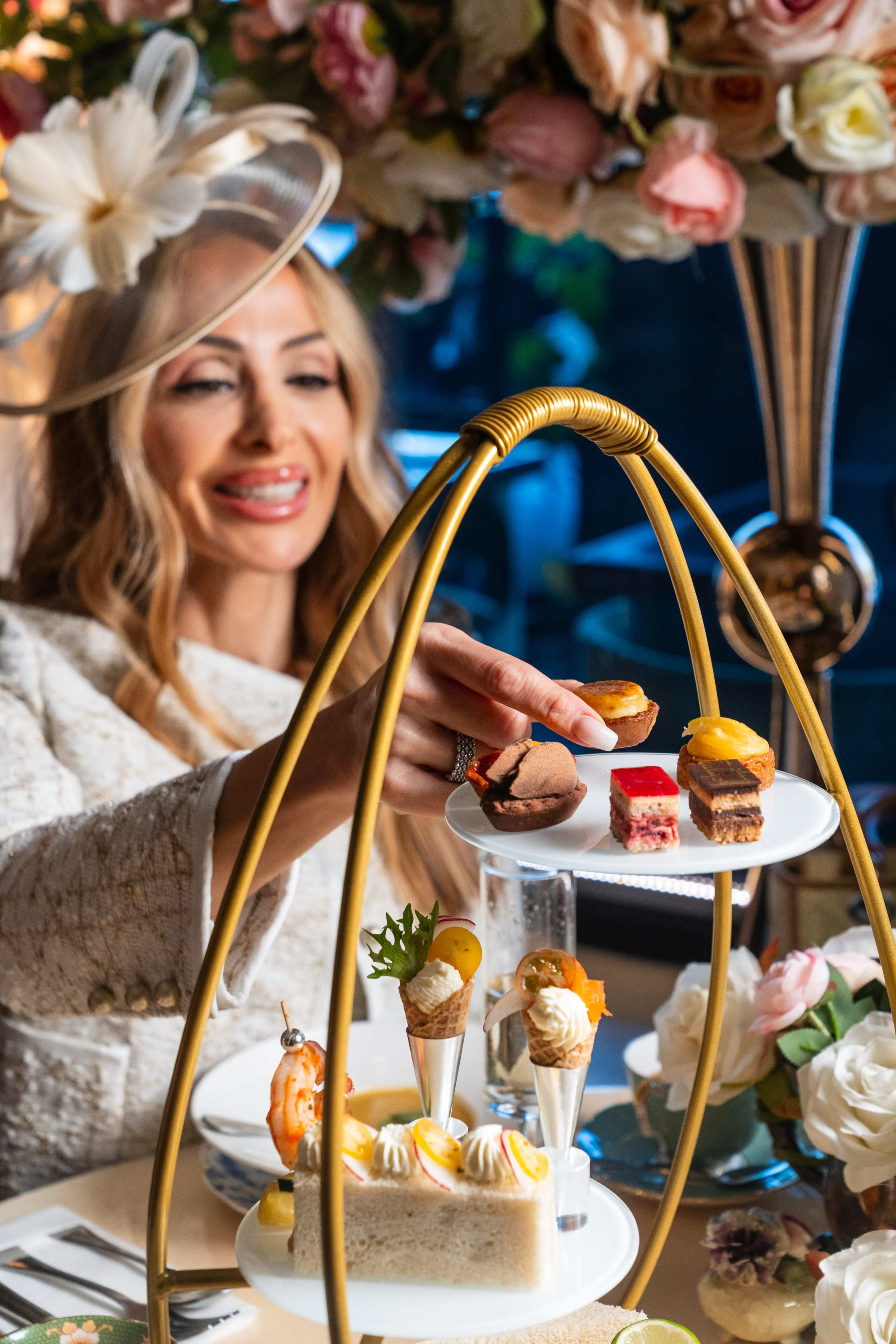 A woman in a white outfit places a dessert onto a tiered stand decorated with assorted pastries during a tea event.