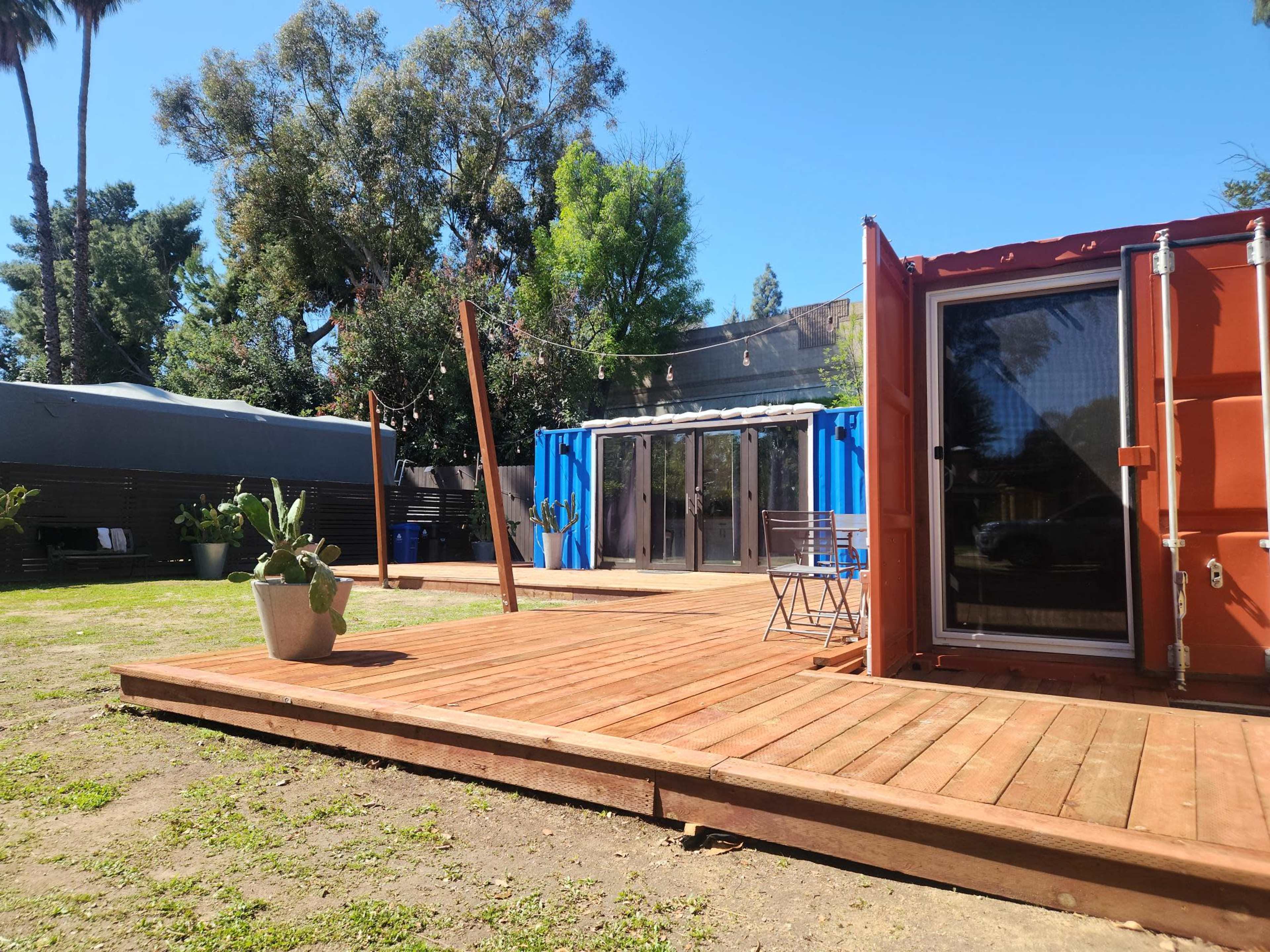 The image shows a wooden deck with two shipping containers converted into a structure, surrounded by greenery and flanked by trees under a clear blue sky.