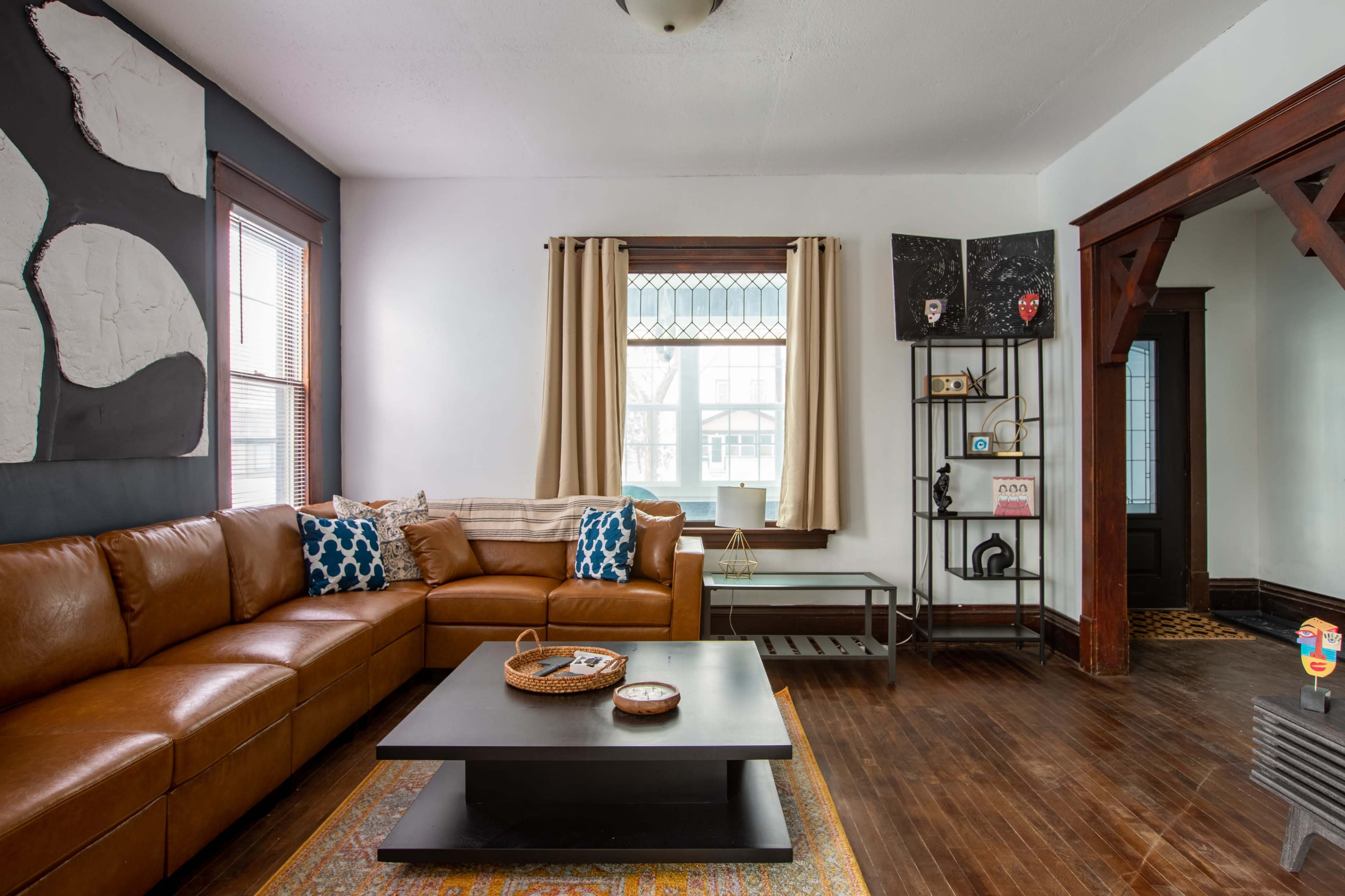 The image shows a living room with a brown sectional sofa, a dark wooden coffee table, and large windows adorned with cream-colored curtains.