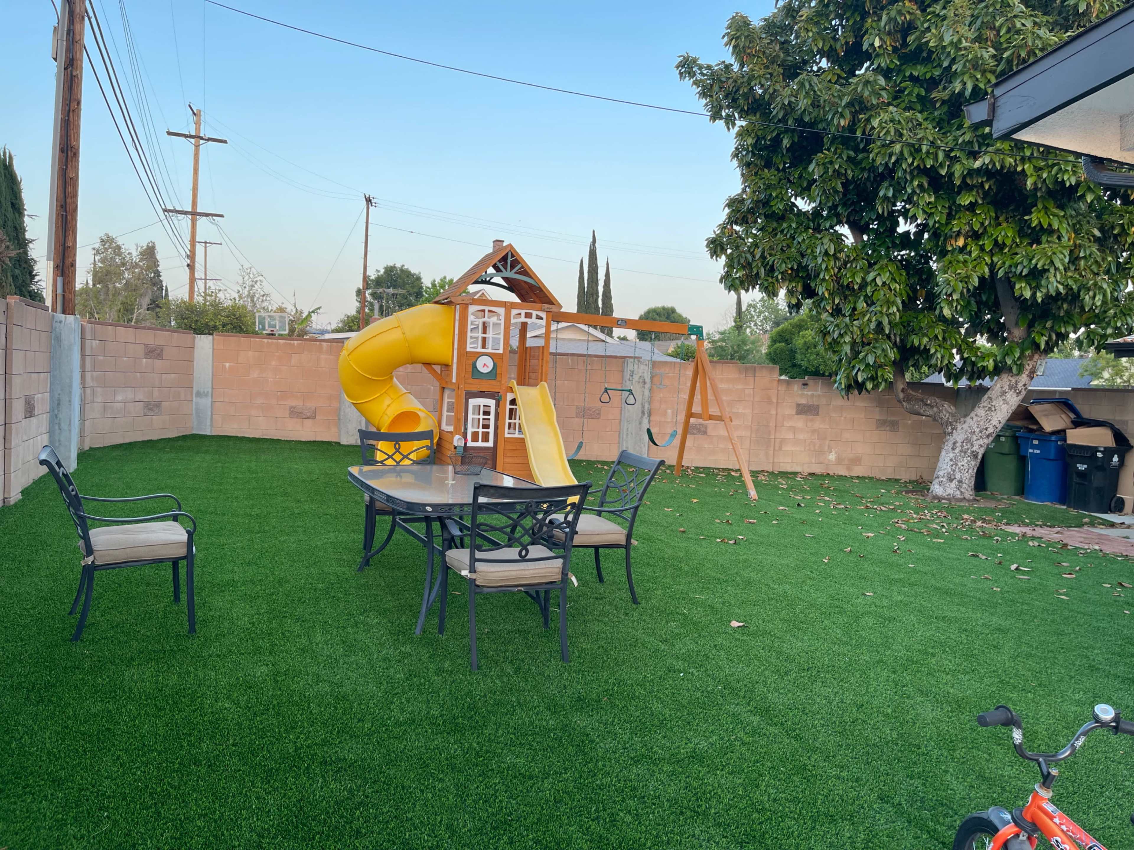 A backyard featuring a yellow slide structure, a swing set, a table with chairs, and artificial grass.