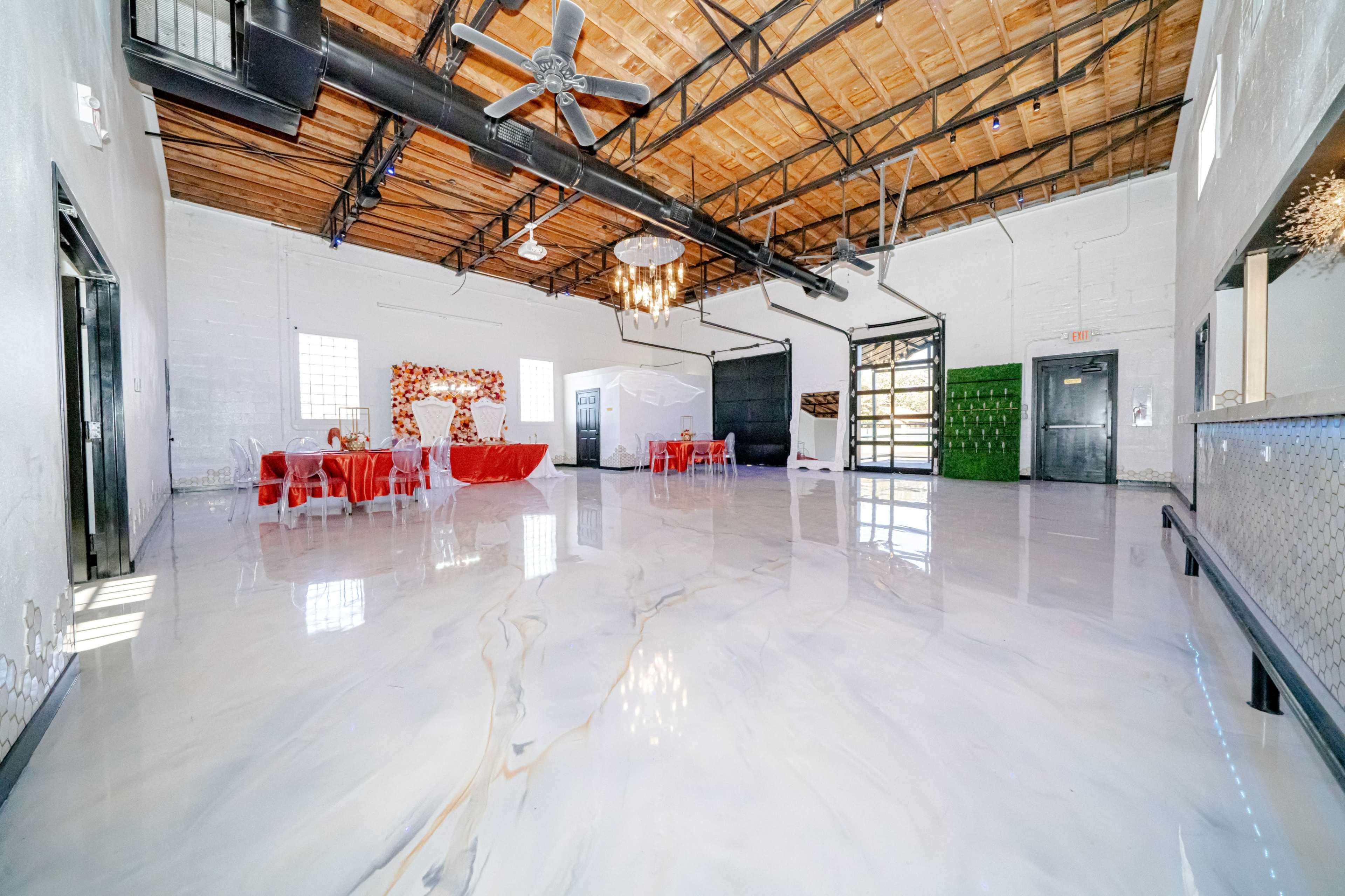 The image shows a spacious, empty event venue with a polished marble floor, exposed wooden beams, and tables set with red tablecloths.