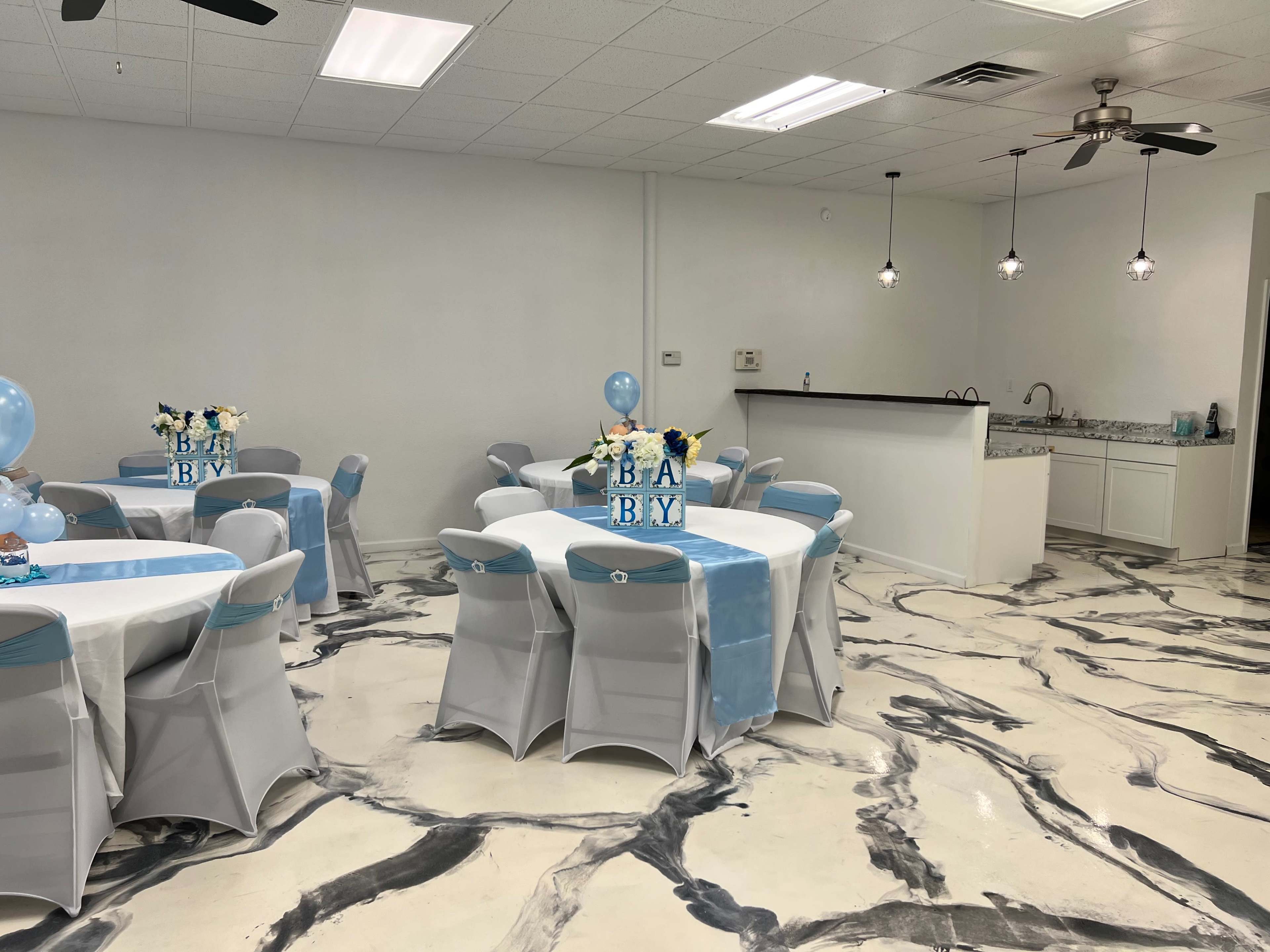 A decorated event space with gray and white marble flooring, round tables covered in white tablecloths with blue accents, and a serving area in the background.
