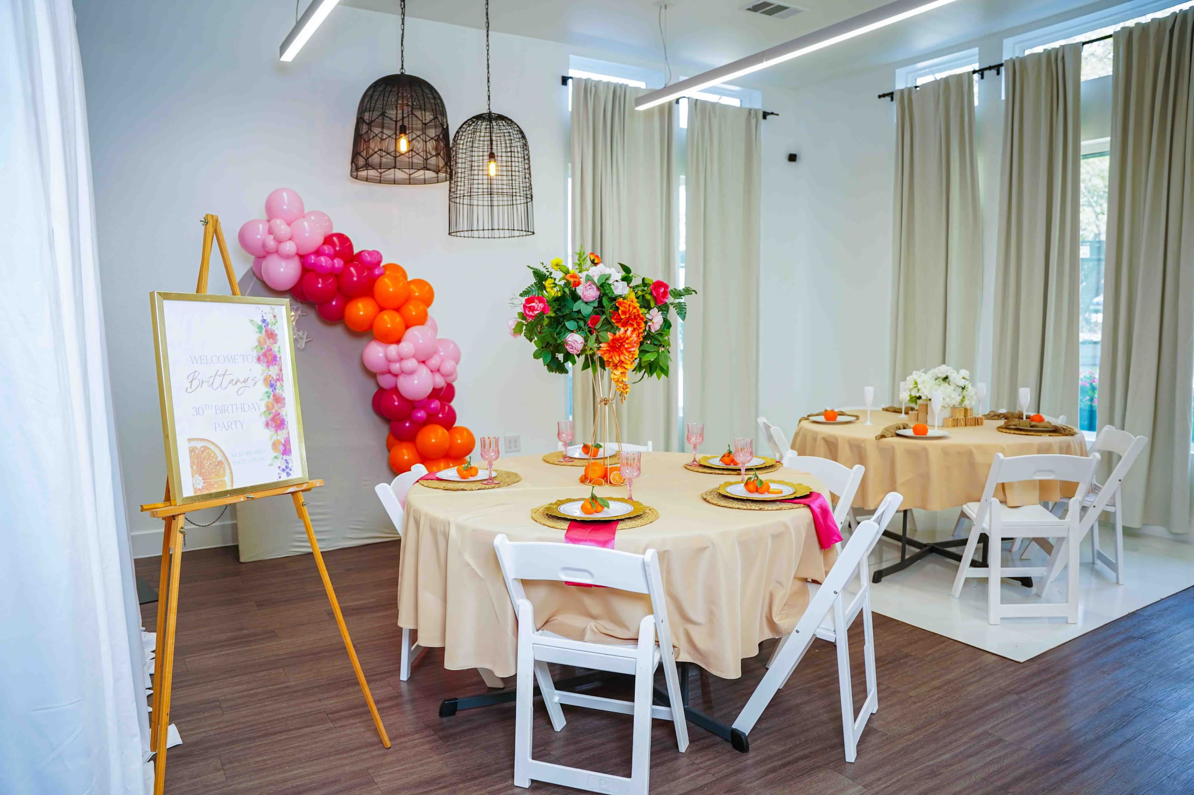 The image shows a decorated dining area with two round tables set for a party, featuring floral centerpieces, colorful balloons in an arch, and a sign welcoming guests.