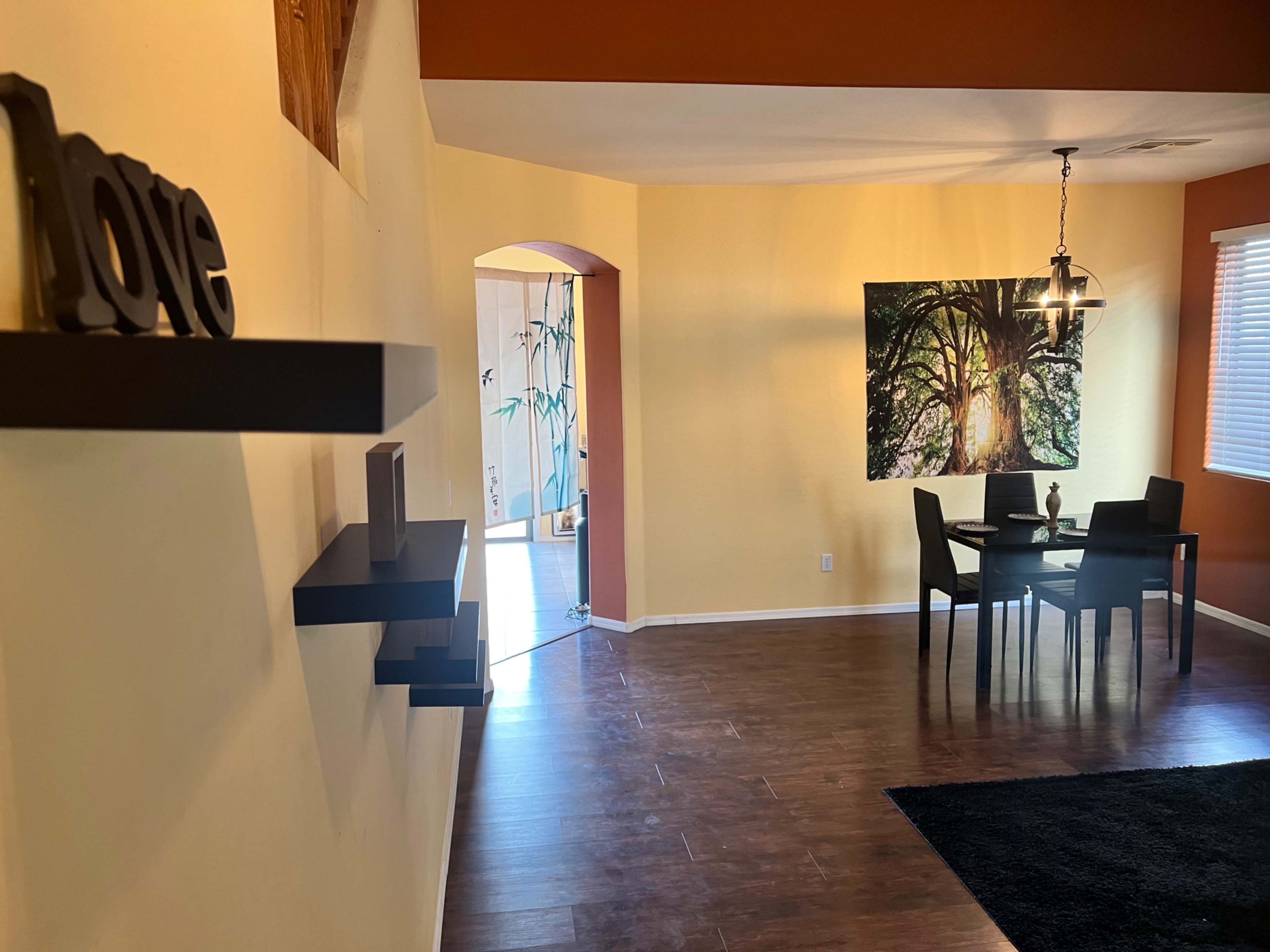 A modern living space with wooden flooring, a small dining table set, a large tree photograph on the wall, and decorative shelves.