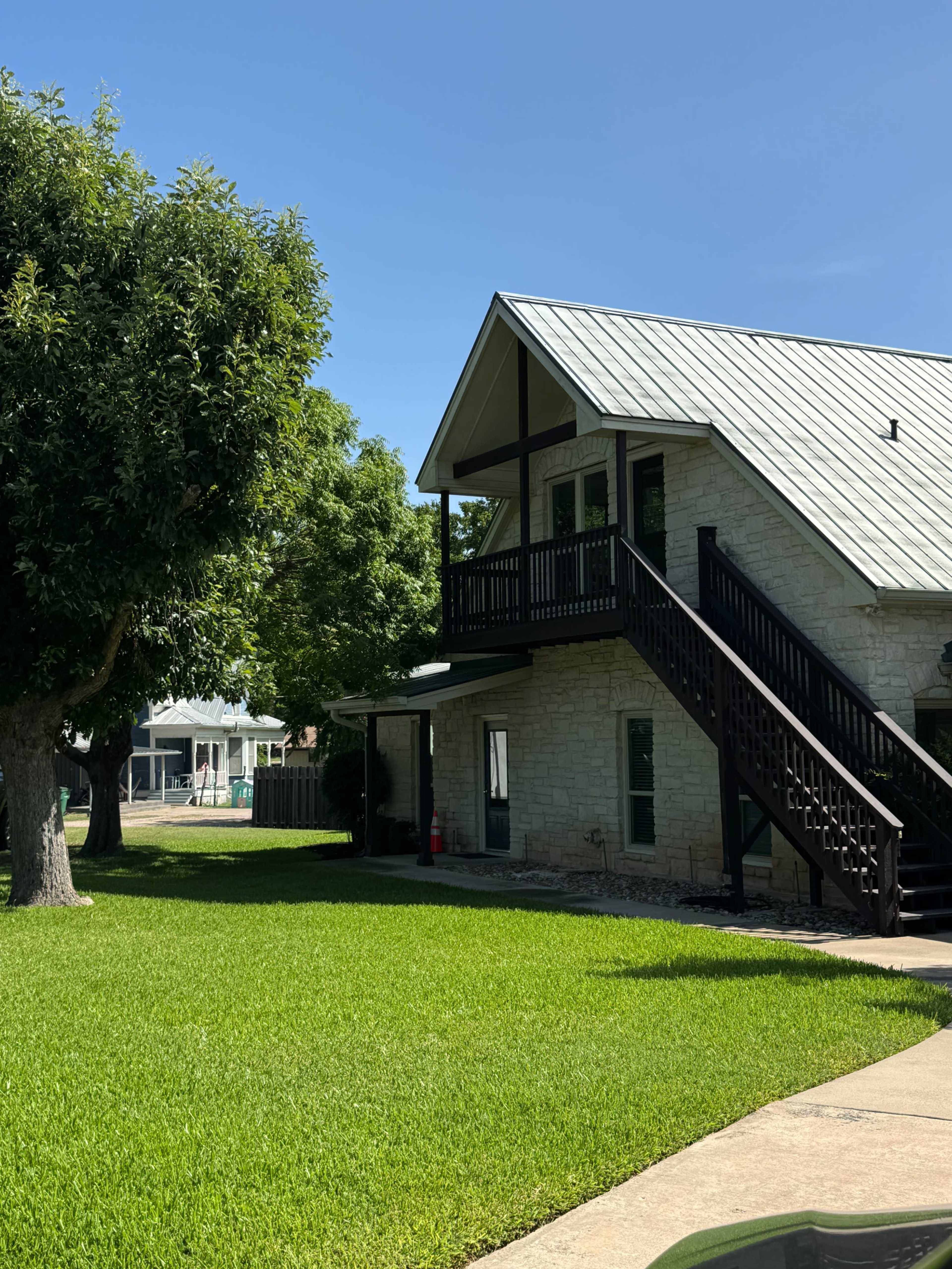 A two-story stone house with a metal roof and a wooden staircase, surrounded by green grass and trees.