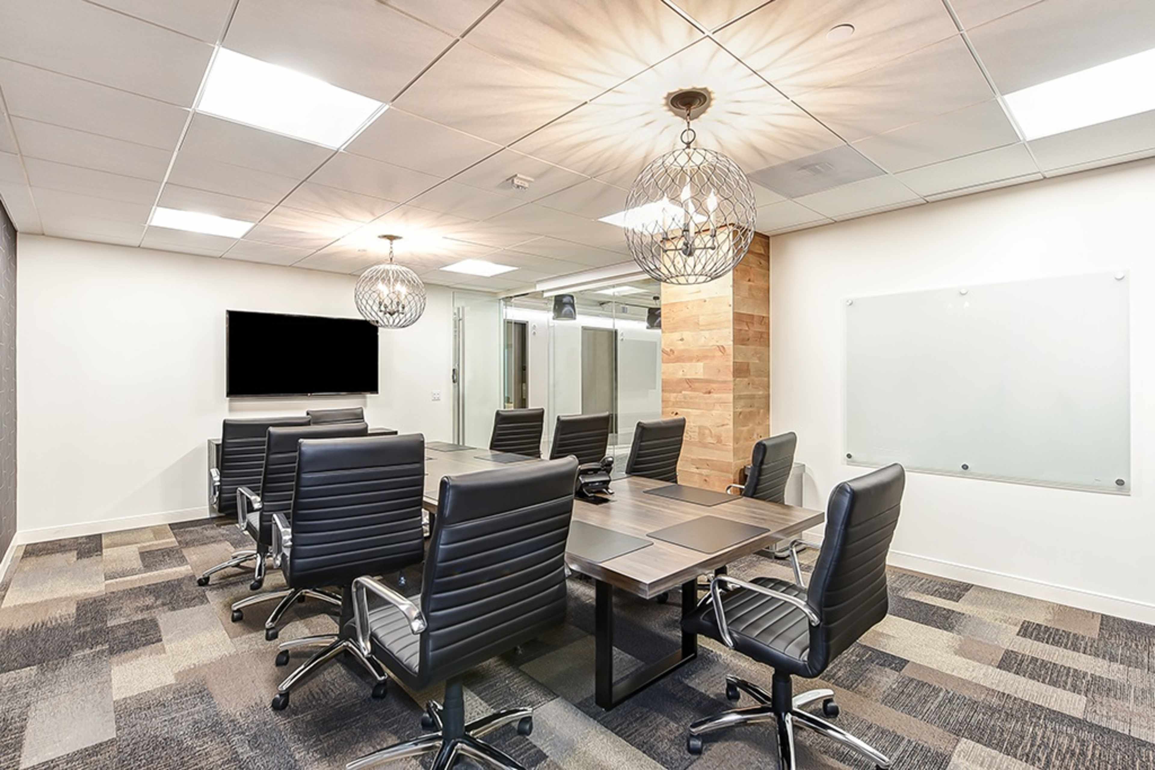 A modern conference room with a large table surrounded by black leather chairs, featuring a wall-mounted screen and two decorative light fixtures.