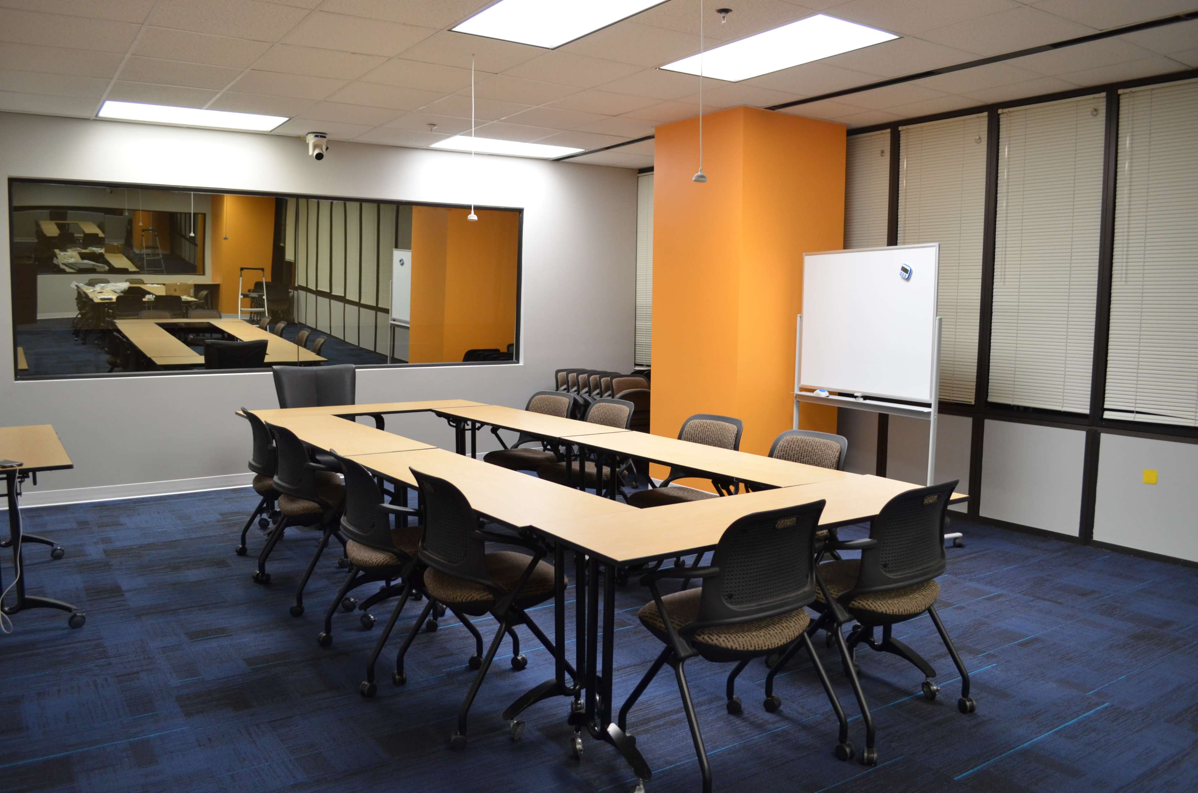The image shows a conference room with a rectangular table surrounded by chairs, a whiteboard, and large windows.