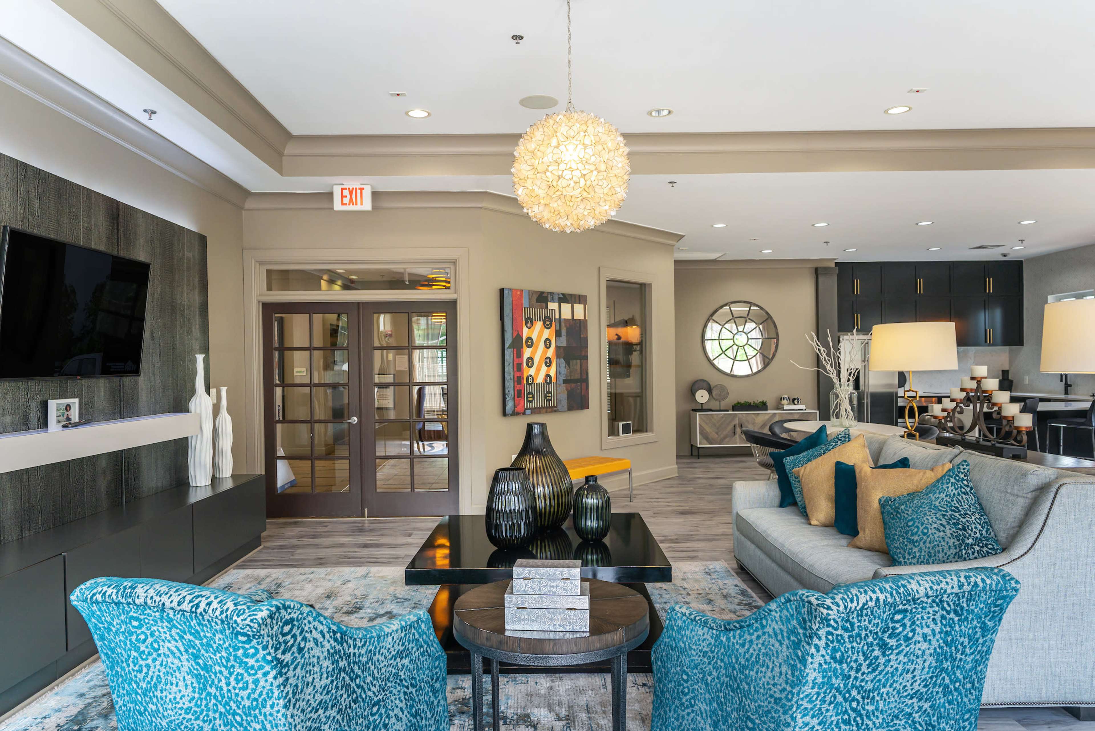 A modern, stylish lounge area featuring a seating arrangement with teal-patterned chairs, a central coffee table, decorative vases, and a large circular window.