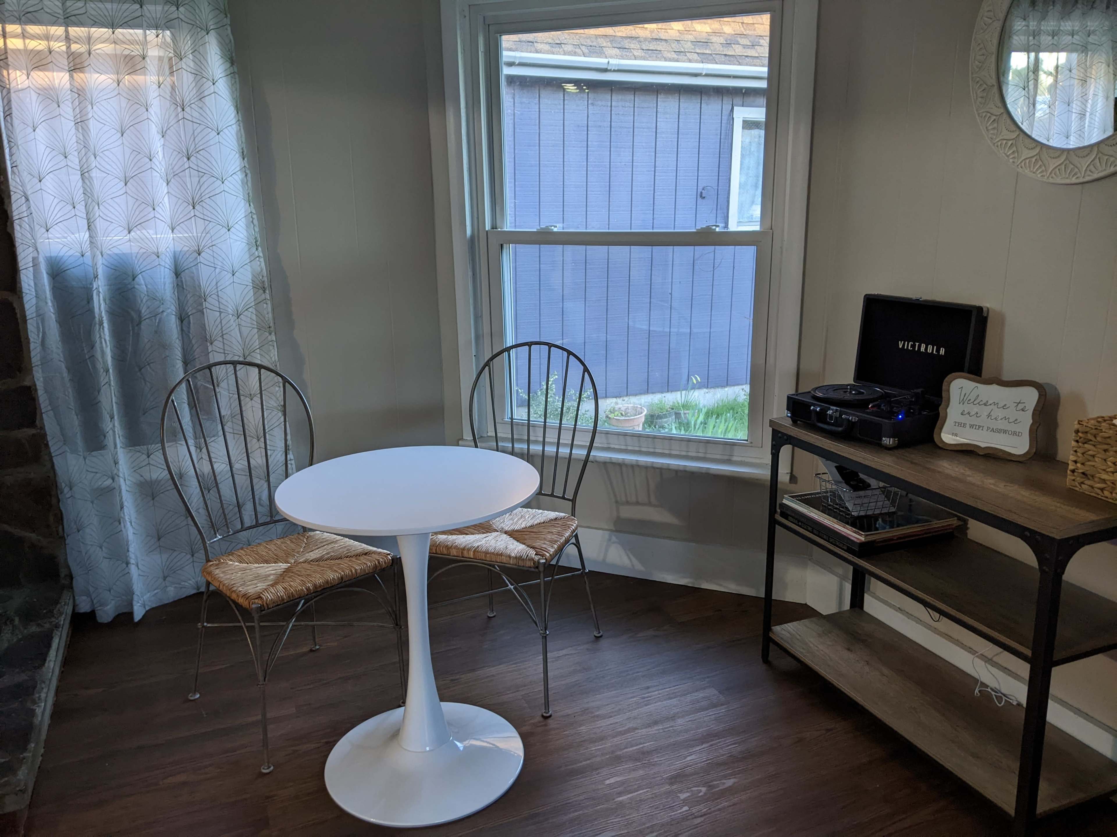 A small dining area features a round table with two wire chairs alongside a shelf with a record player and decorative items, all set near a window.