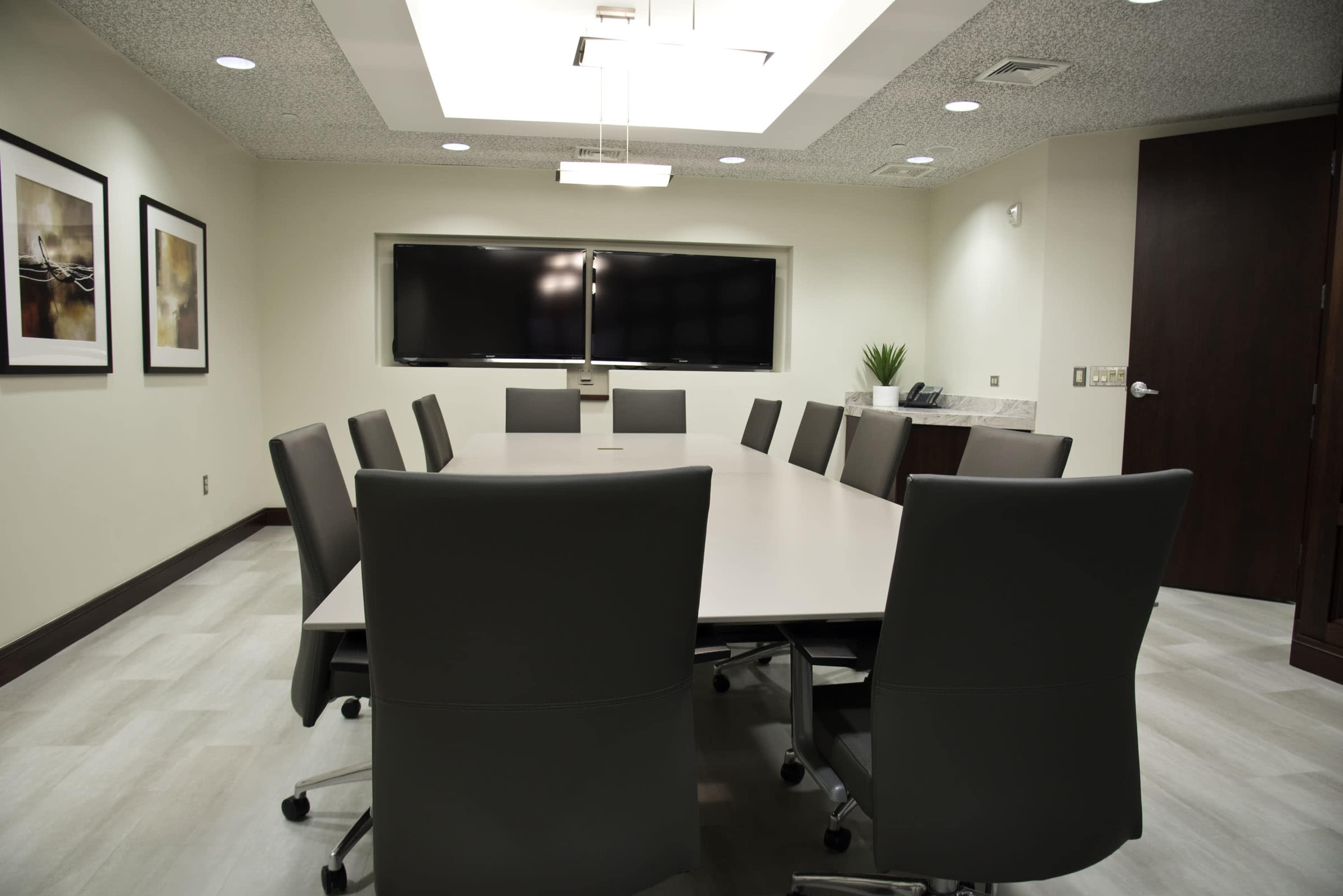 A modern conference room features a rectangular table surrounded by ten gray chairs, with two large screens mounted on the wall and framed art on the adjacent walls.