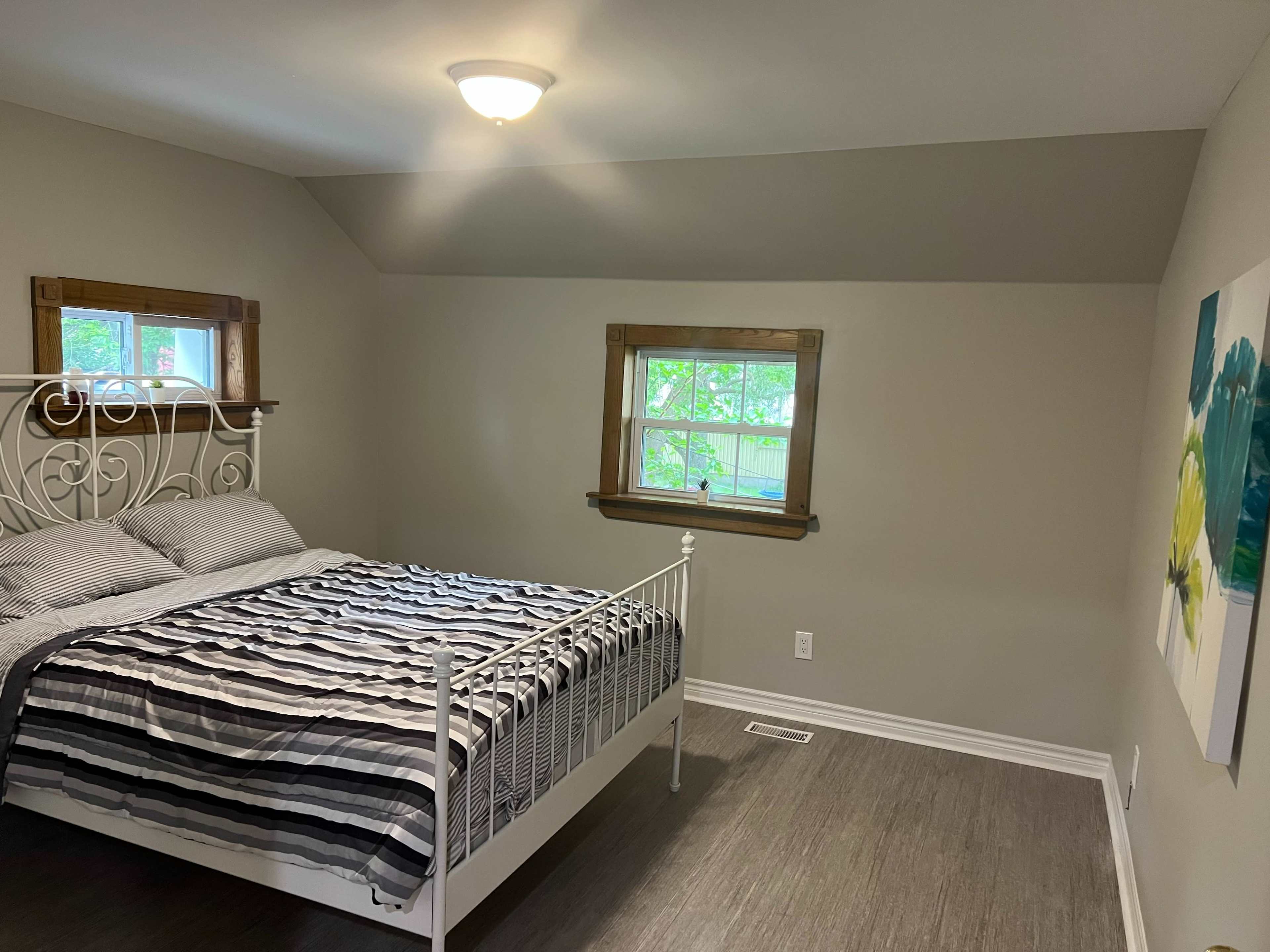 The image shows a neatly arranged bedroom with a white metal bed, striped bedding, and two windows allowing natural light.