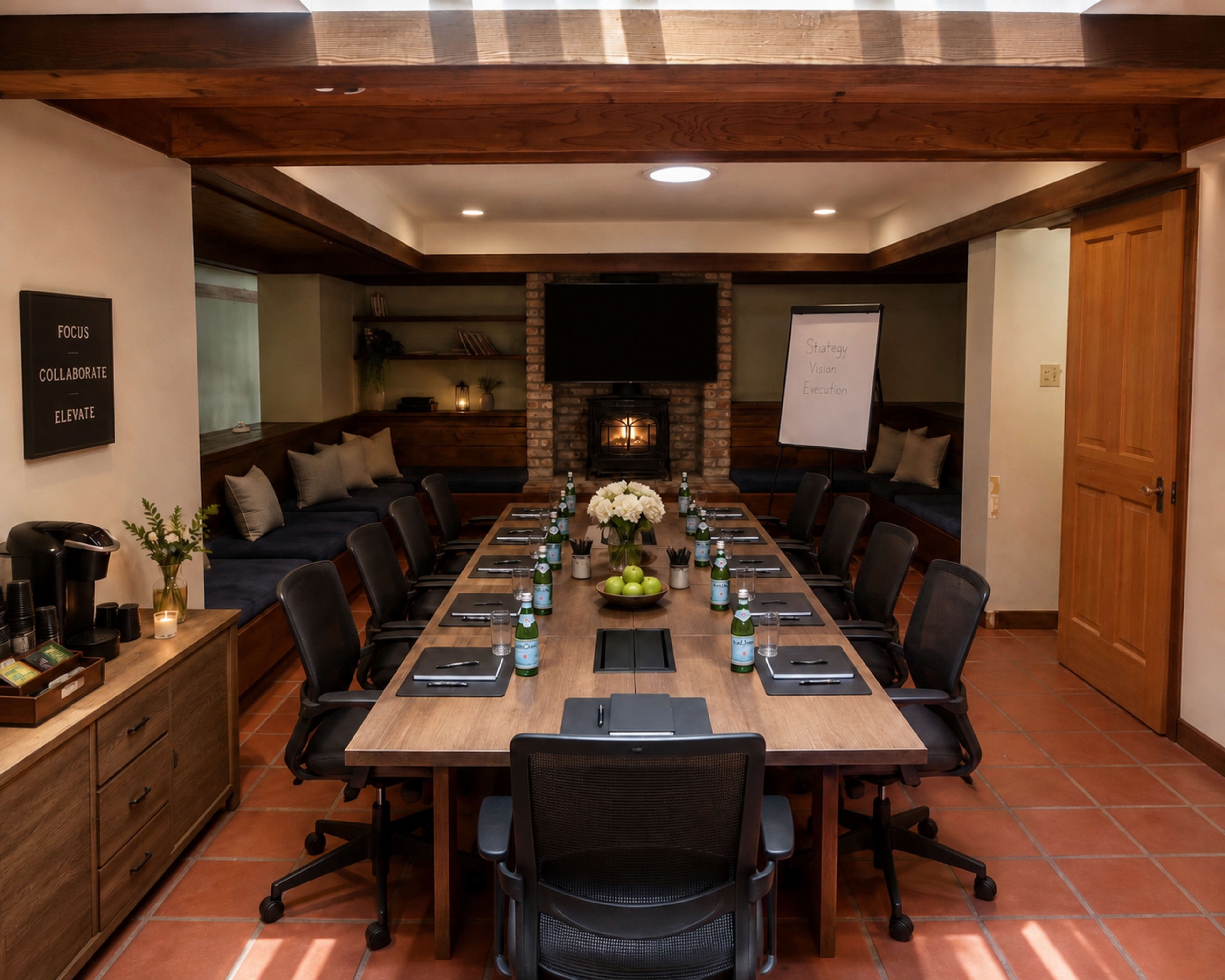 A long conference table surrounded by chairs is set up in a spacious meeting room with a fireplace, large screen, and natural light.
