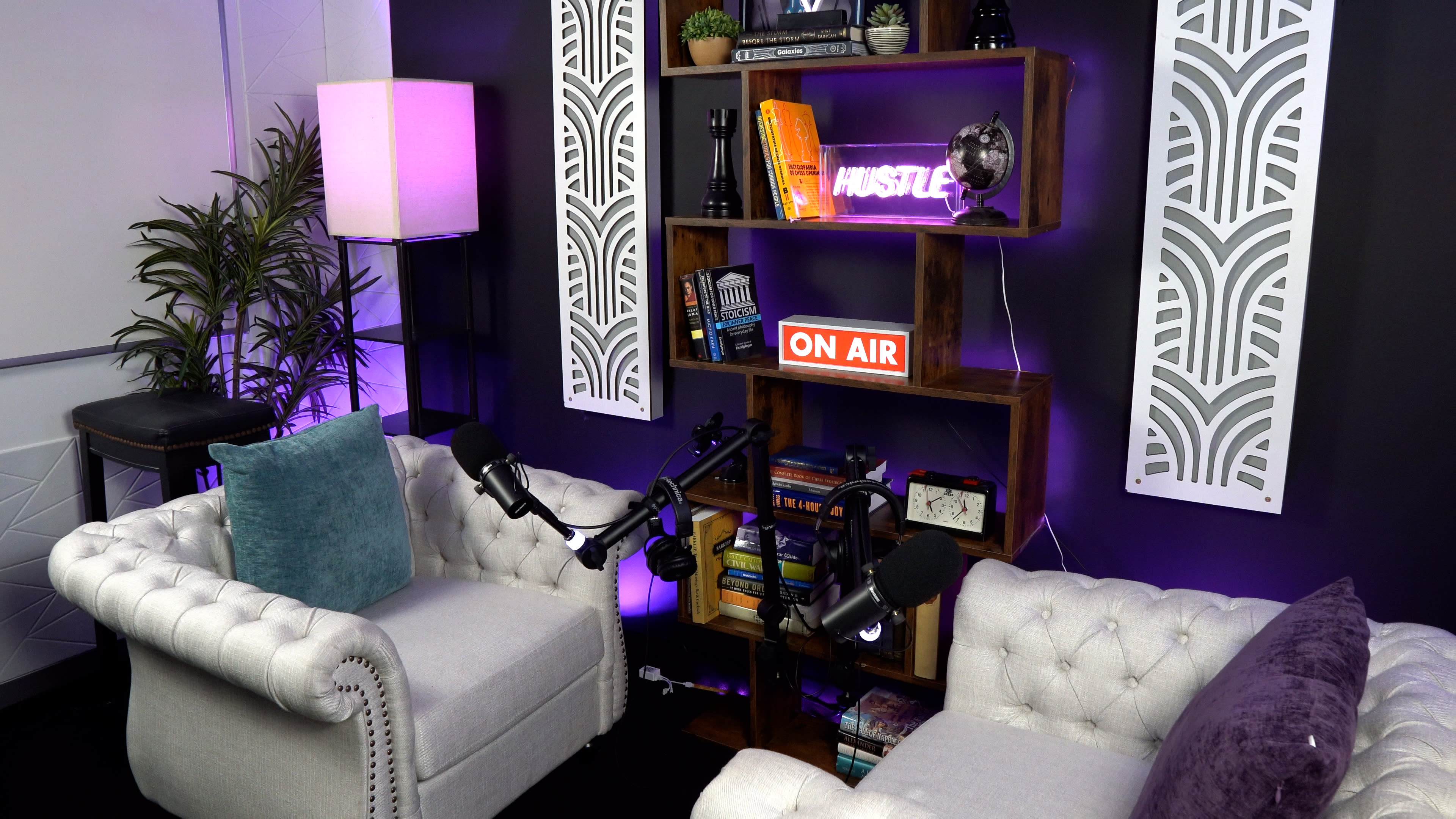 A small podcasting studio with two tufted armchairs facing a bookshelf, illuminated by neon signs and soft lighting.
