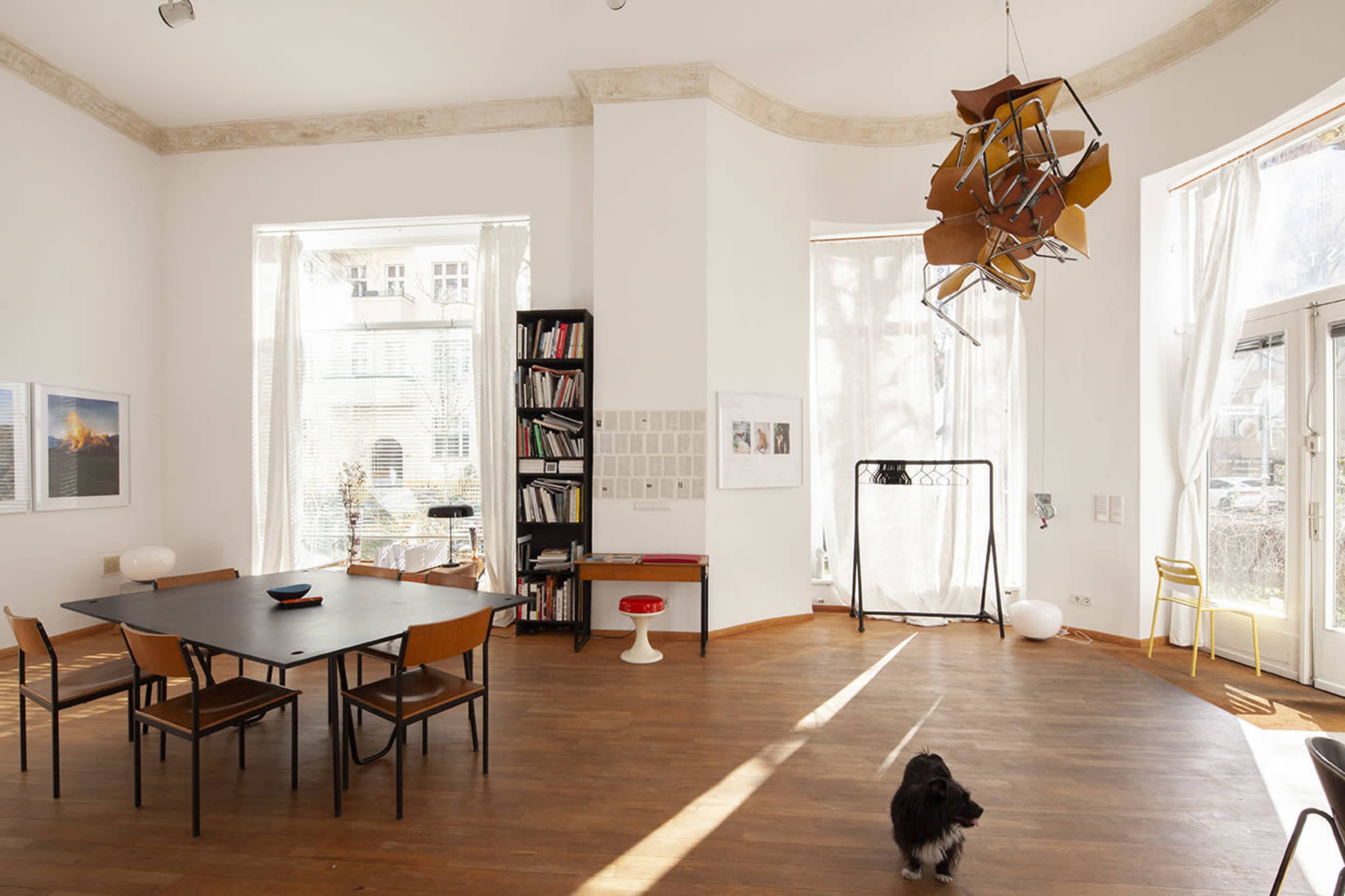 A spacious room with a wooden floor, a rectangular black dining table surrounded by chairs, bookshelves, and large windows allowing natural light to enter.