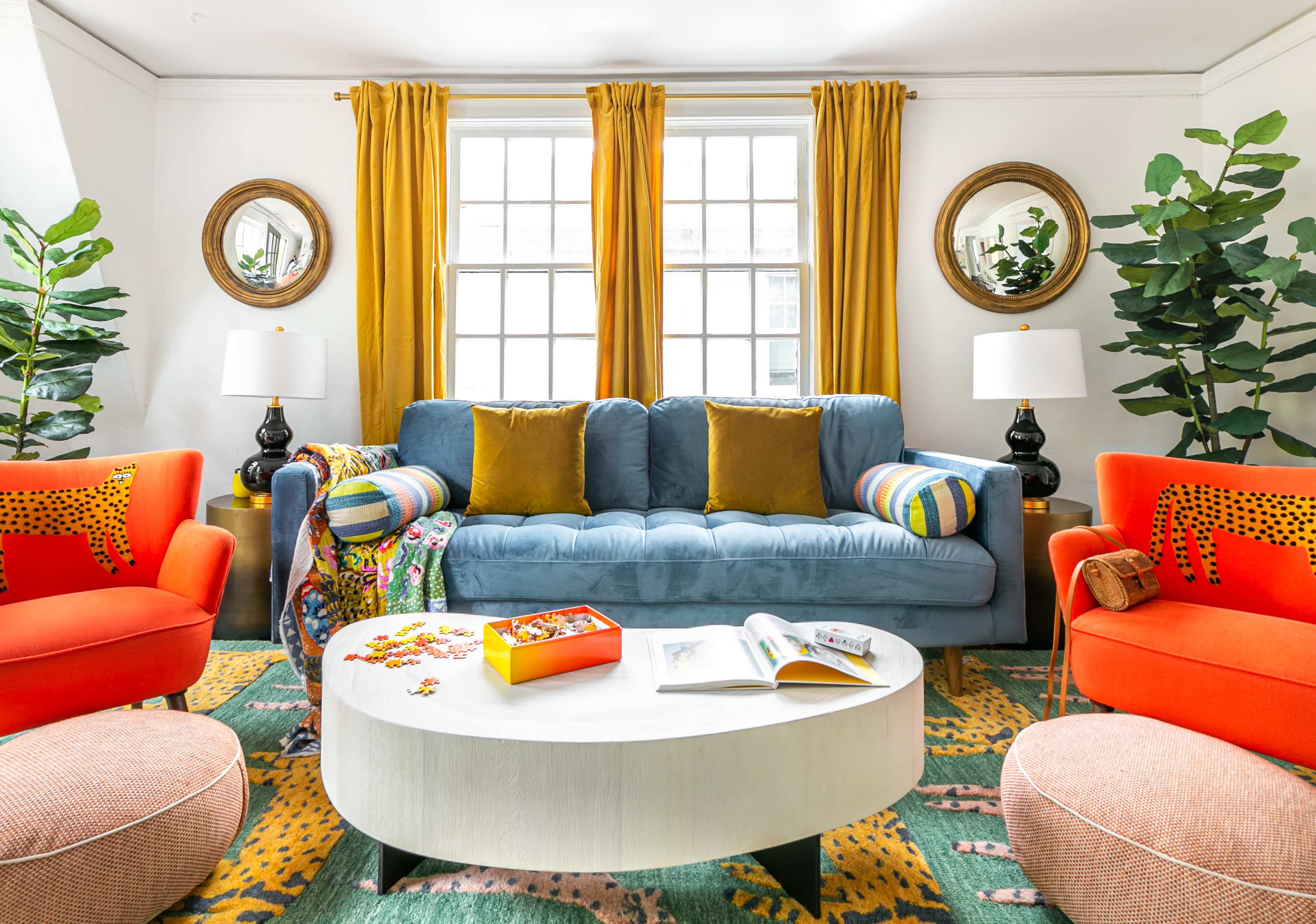 A colorful living room featuring a blue sofa, orange armchairs, a wooden coffee table, and plants, with large windows adorned with yellow curtains.