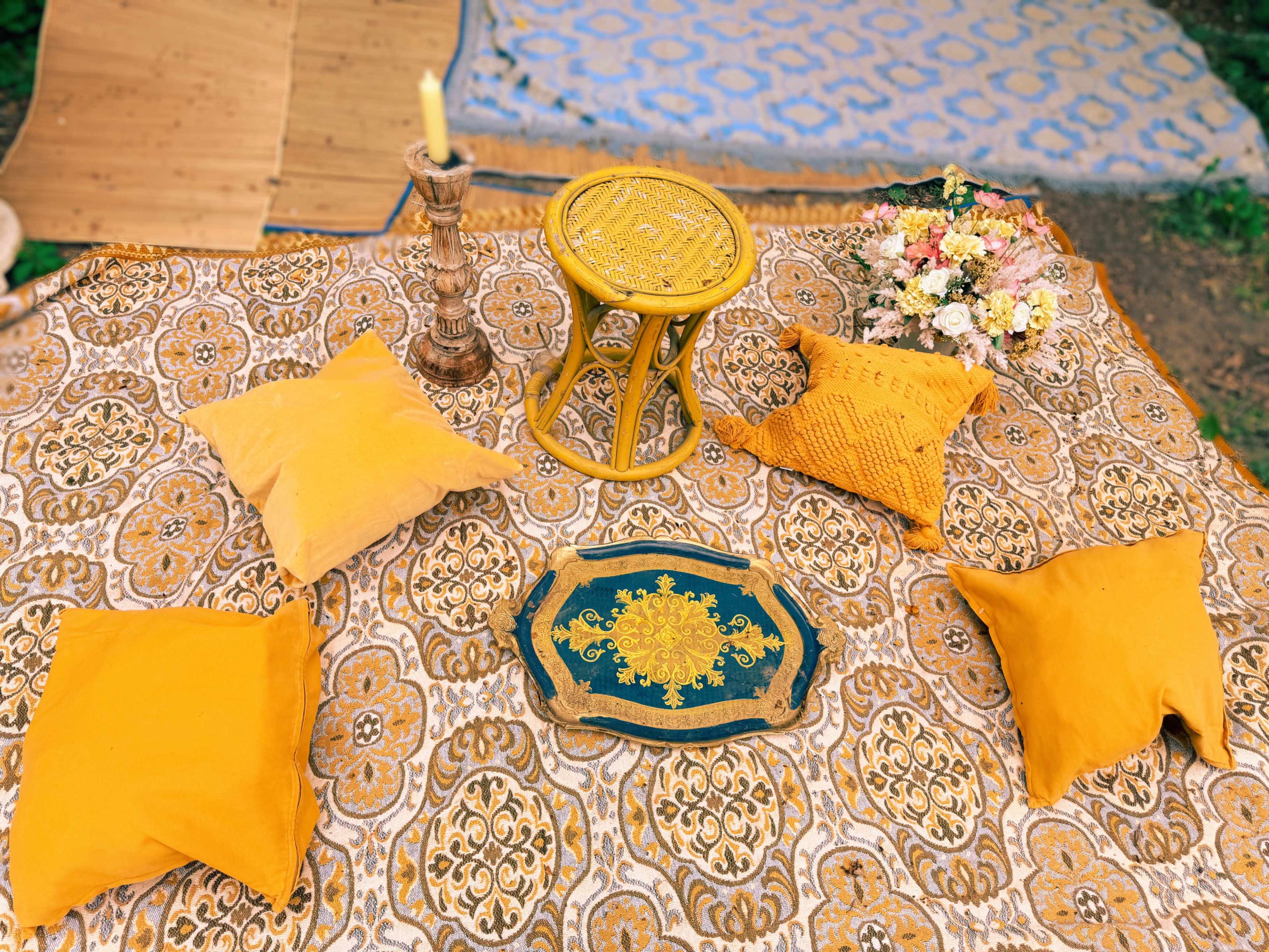 A table set with decorative items and yellow pillows, surrounded by a patterned fabric in an outdoor setting.